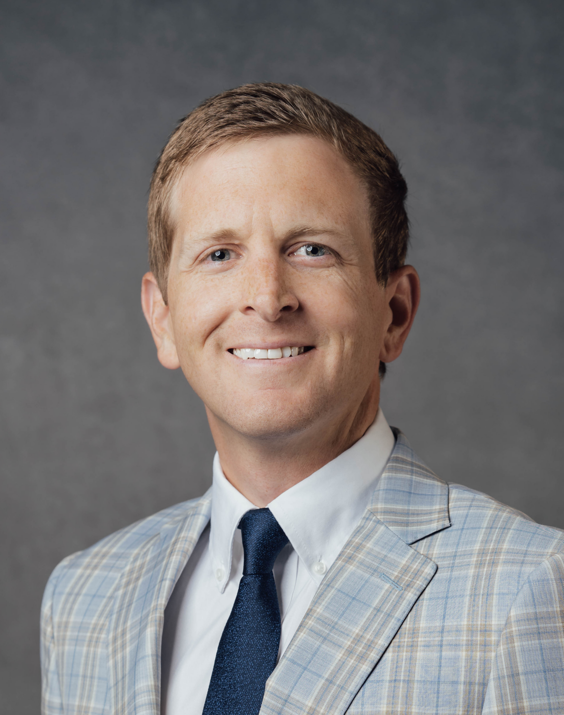 Studio headshot of a smiling person in a light grey and blue suit and navy tie on a dark grey background