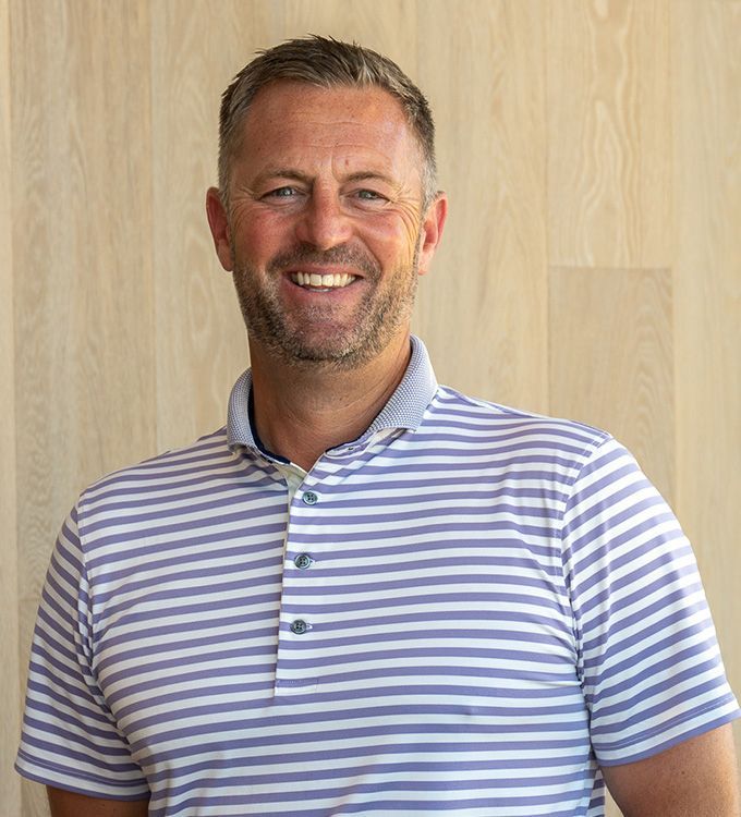 Smiling man in a blue-and-white striped polo shirt standing against a light wood wall