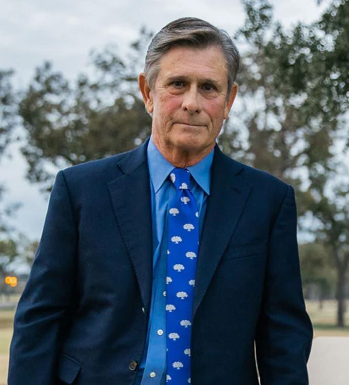 Man in a navy suit and blue tie standing outdoors on a tree-lined path