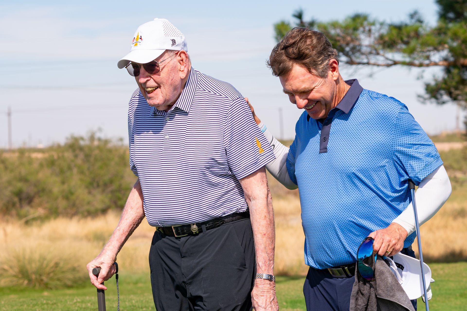Two golfers walking on a sunny course, one in a white cap and striped shirt, the other in a blue shirt with a white sleeve.