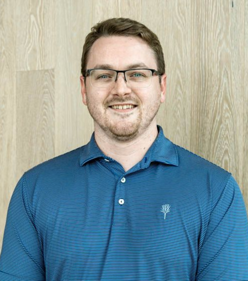 Smiling person in glasses wearing a blue polo shirt against a light wood wall