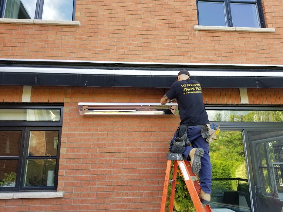 A man is standing on a ladder fixing an awning on the side of a brick building.