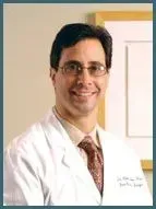 A doctor with dark hair and glasses wears a white lab coat over a dress shirt and tie, smiling against a neutral wall.