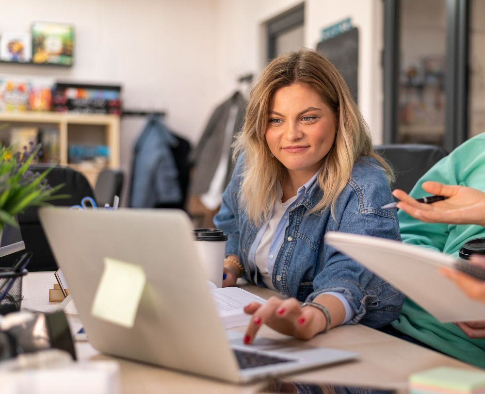 Vrouw in spijkerjasje, werkend op een laptop aan een bureau, met een geconcentreerde blik naar het scherm.