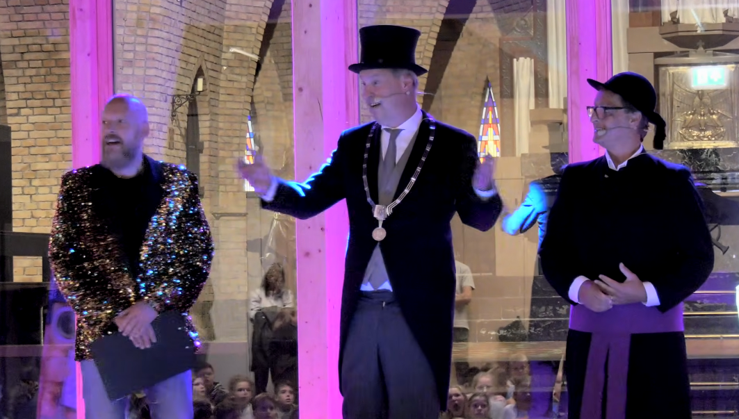 Drie personen in galakleding staan ​​op een verlicht podium, van wie er één een hoge hoed draagt ​​en een wandelstokachtig voorwerp vasthoudt.