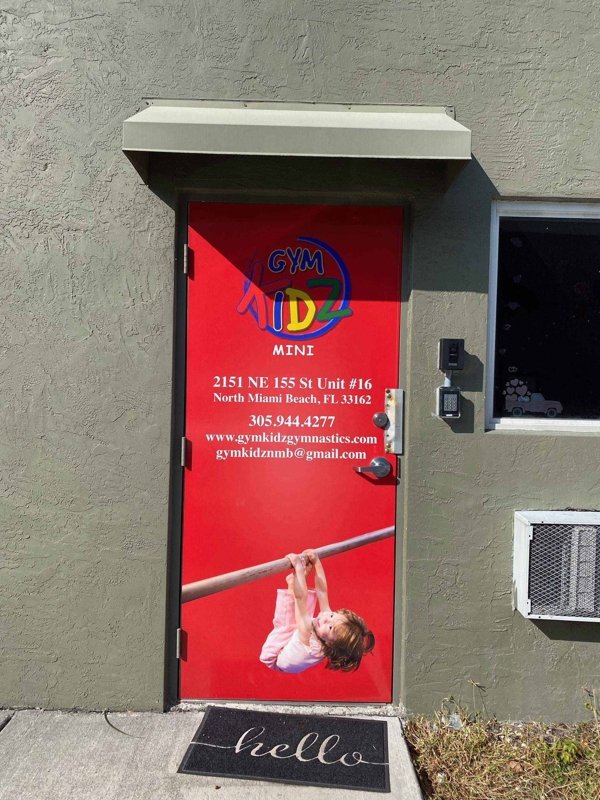 Red door with Hindu poster and hanging baby image, set in a gray wall exterior.