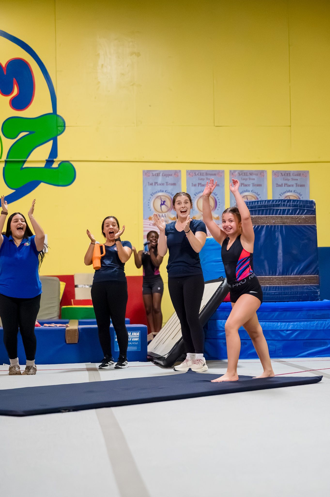 Gymnastics class posing on a mat indoors, with one child in a pink-and-black leotard in front.