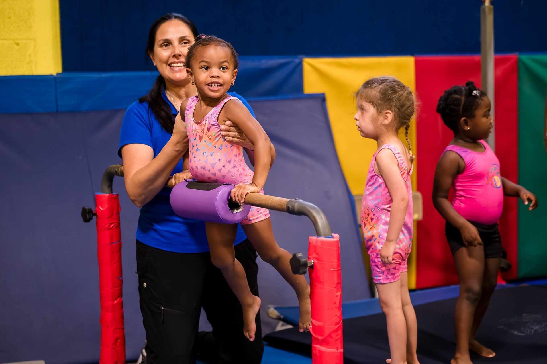 Woman helping a child on a gymnastics bar, with two other children waiting nearby in a gym