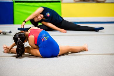 Gymnasts stretching on a mat, one in blue and red in the foreground, another in black in the background.