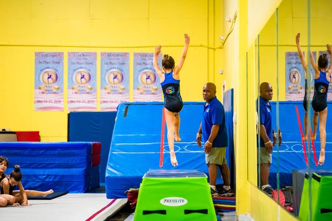 Gymnast mid-jump in a bright gym, reaching for hanging rings near blue mats and a coach.