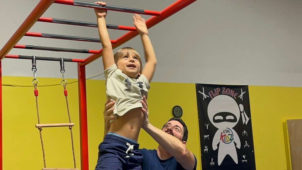 Child hanging from monkey bars in a colorful indoor gym while an adult spots them below