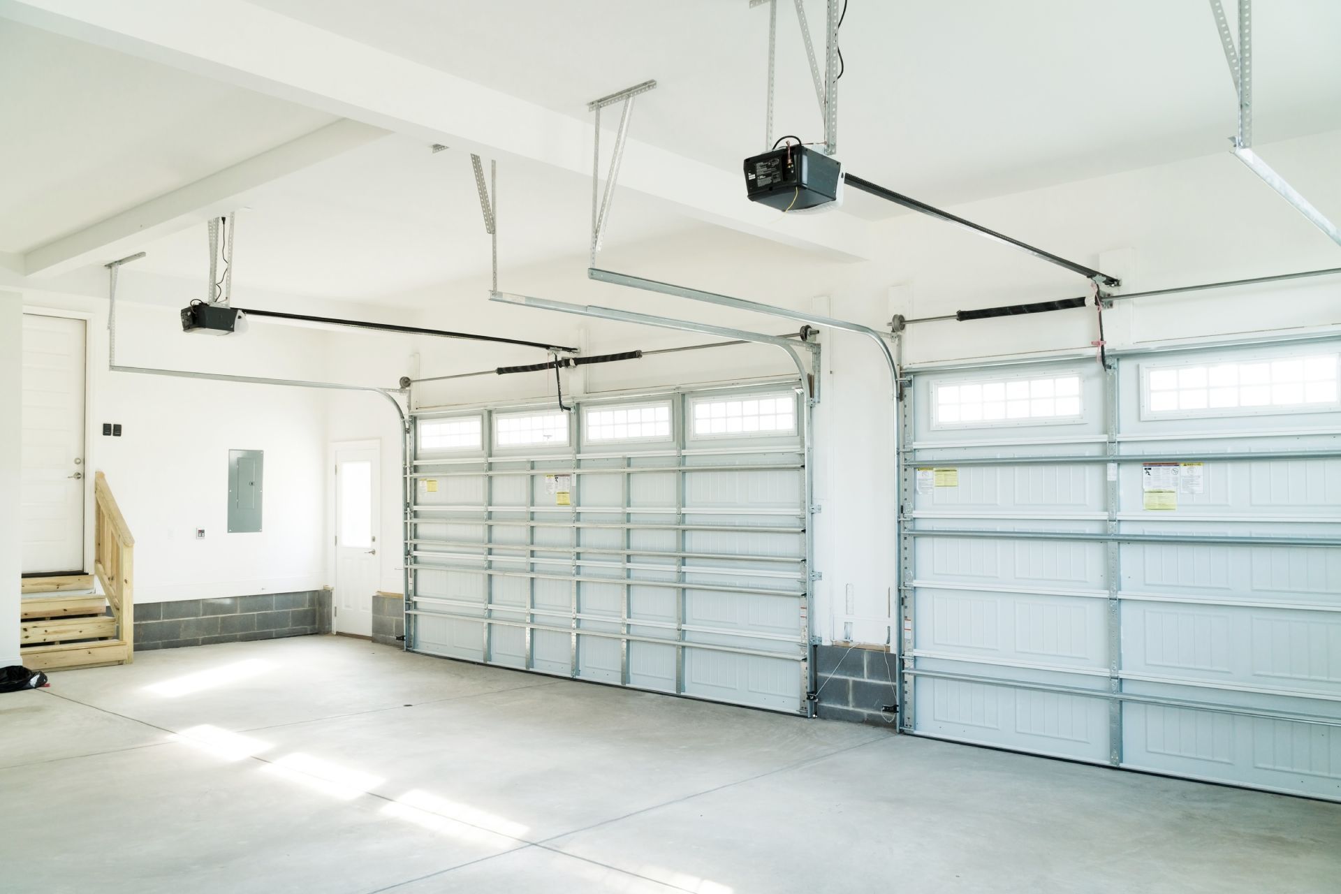 Empty garage with two doors, ceiling-mounted openers, a side door, and unfinished interior details.
