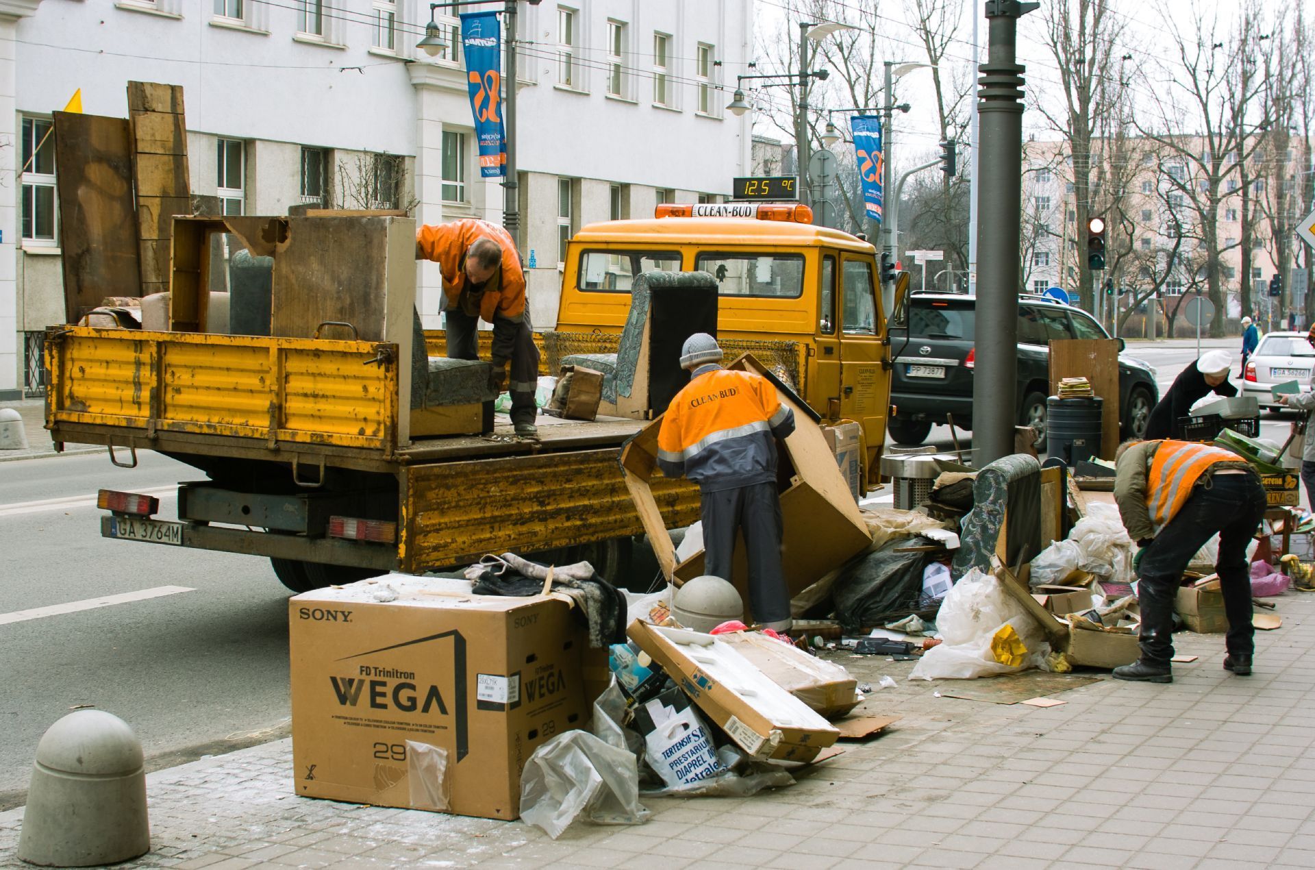 Street cleanup: workers in orange vests loading debris into a yellow truck on a city street.