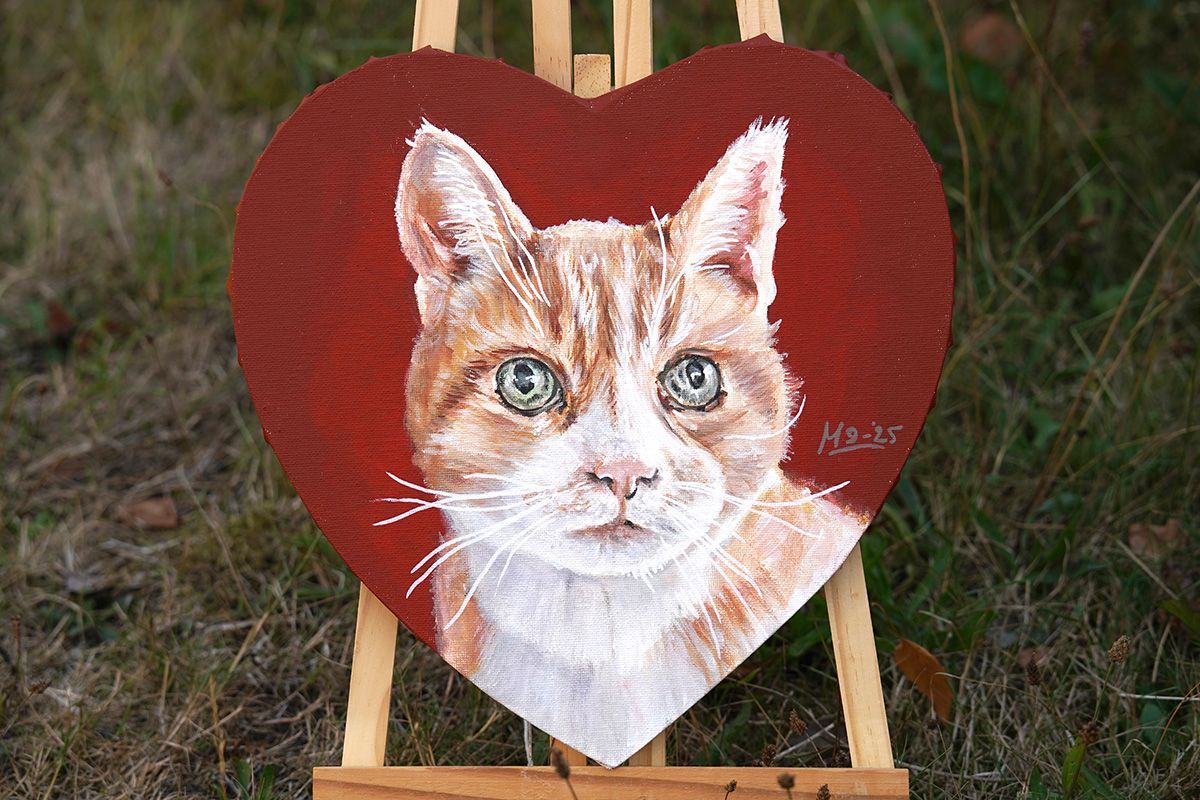 Hartvormig schilderij van een oranje en witte kat met lichtblauwe ogen, op een kleine houten ezel in het gras.