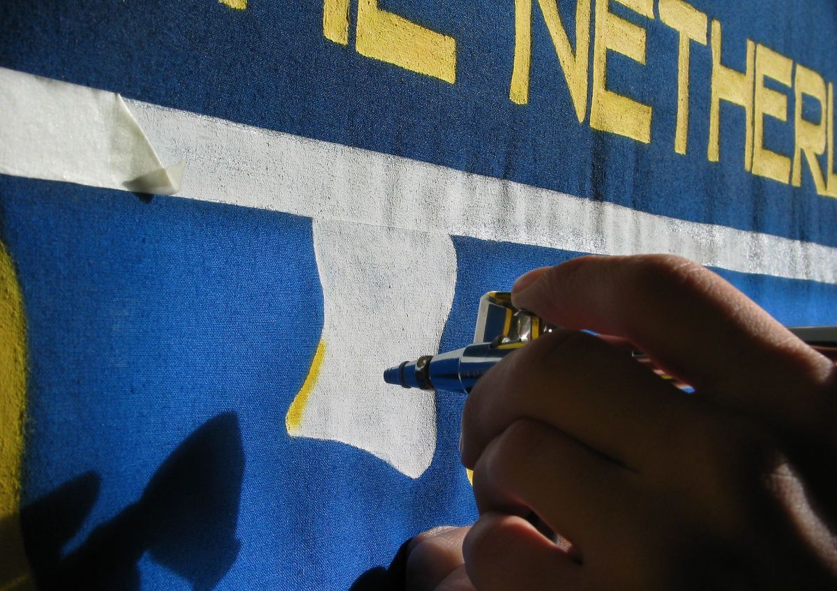 De hand van een persoon gebruikt een stift om een ​​witte vorm op een blauwe stof te sjabloneren. Gele tekst is zichtbaar.
