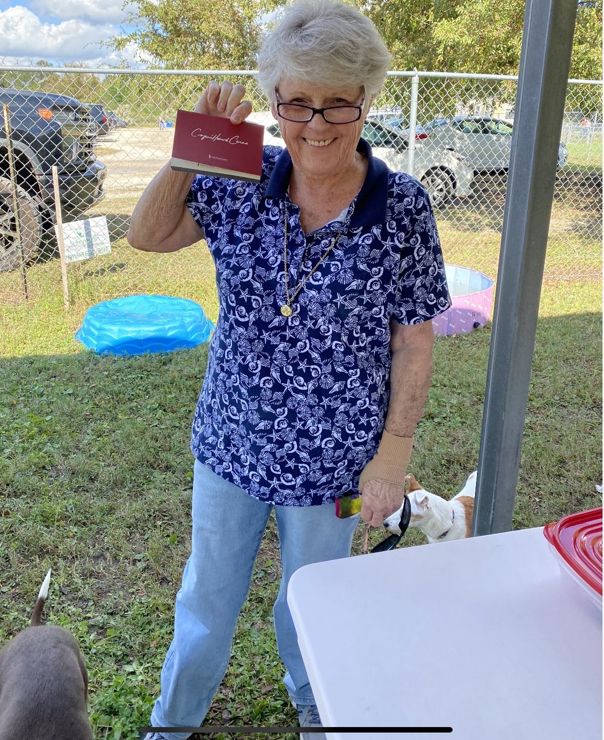 An elderly woman is standing in the grass holding a book.