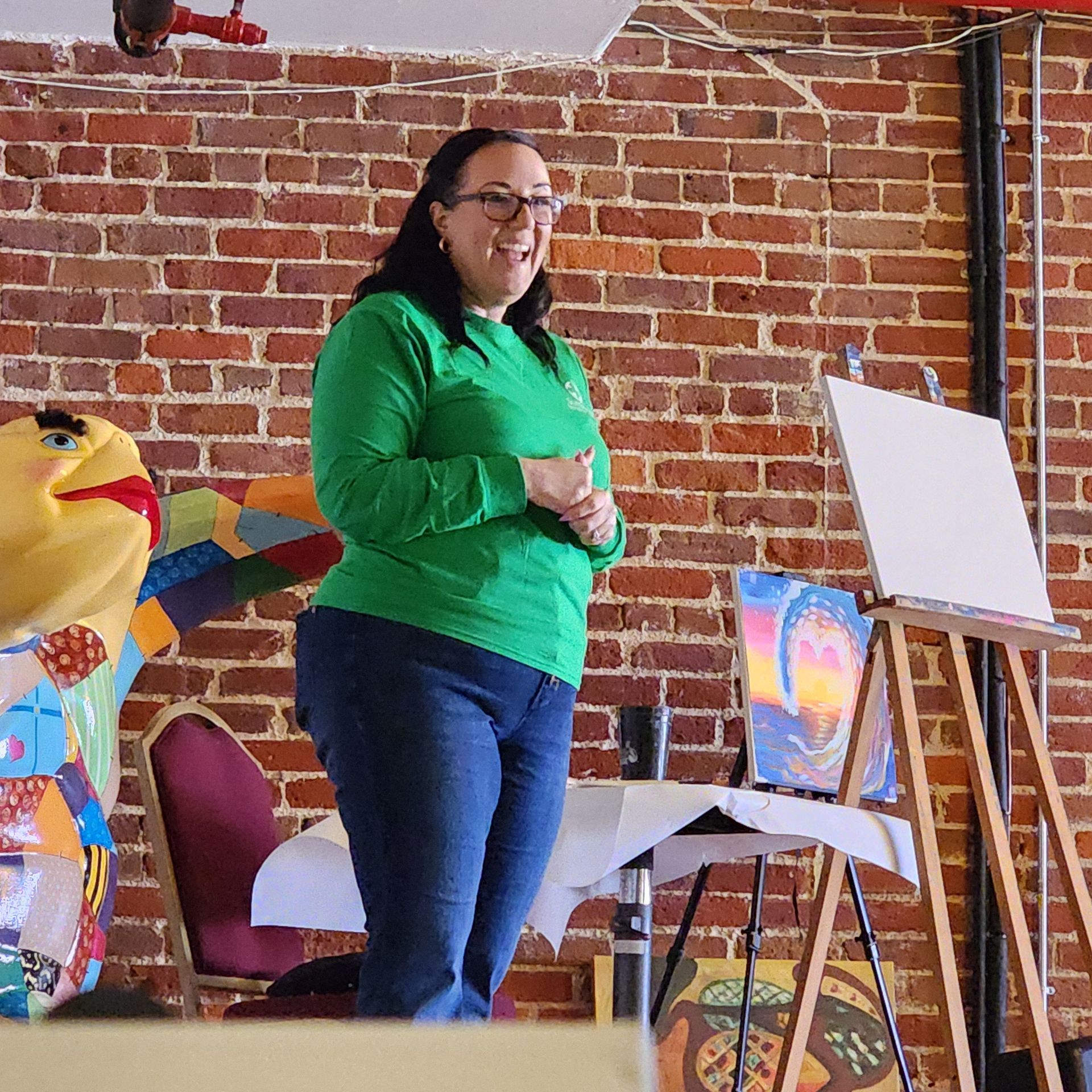 Woman in green sweater stands beside an easel in a brick-walled room, smiling during an art event.