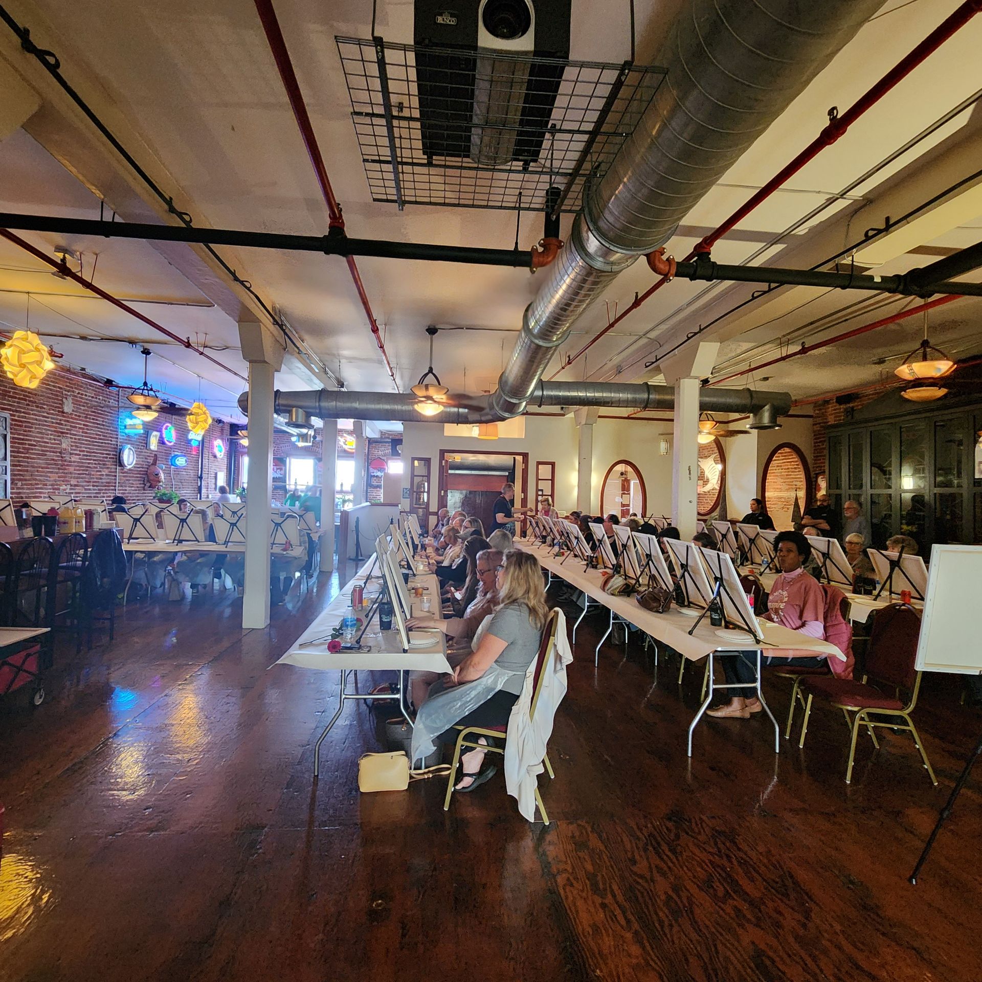 Banquet hall with long dining tables, seated guests, and exposed ceiling ductwork under warm lighting