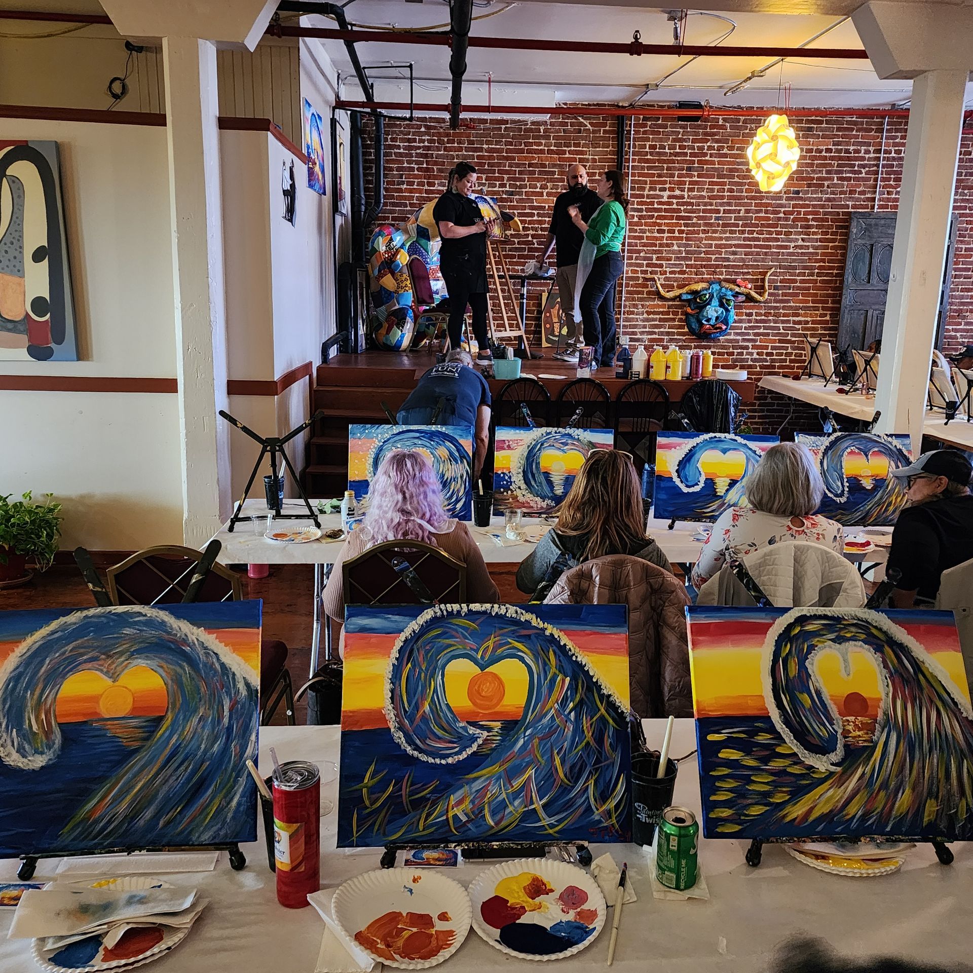 Artists painting heart-themed canvases at a workshop with colorful easels and supplies in front.