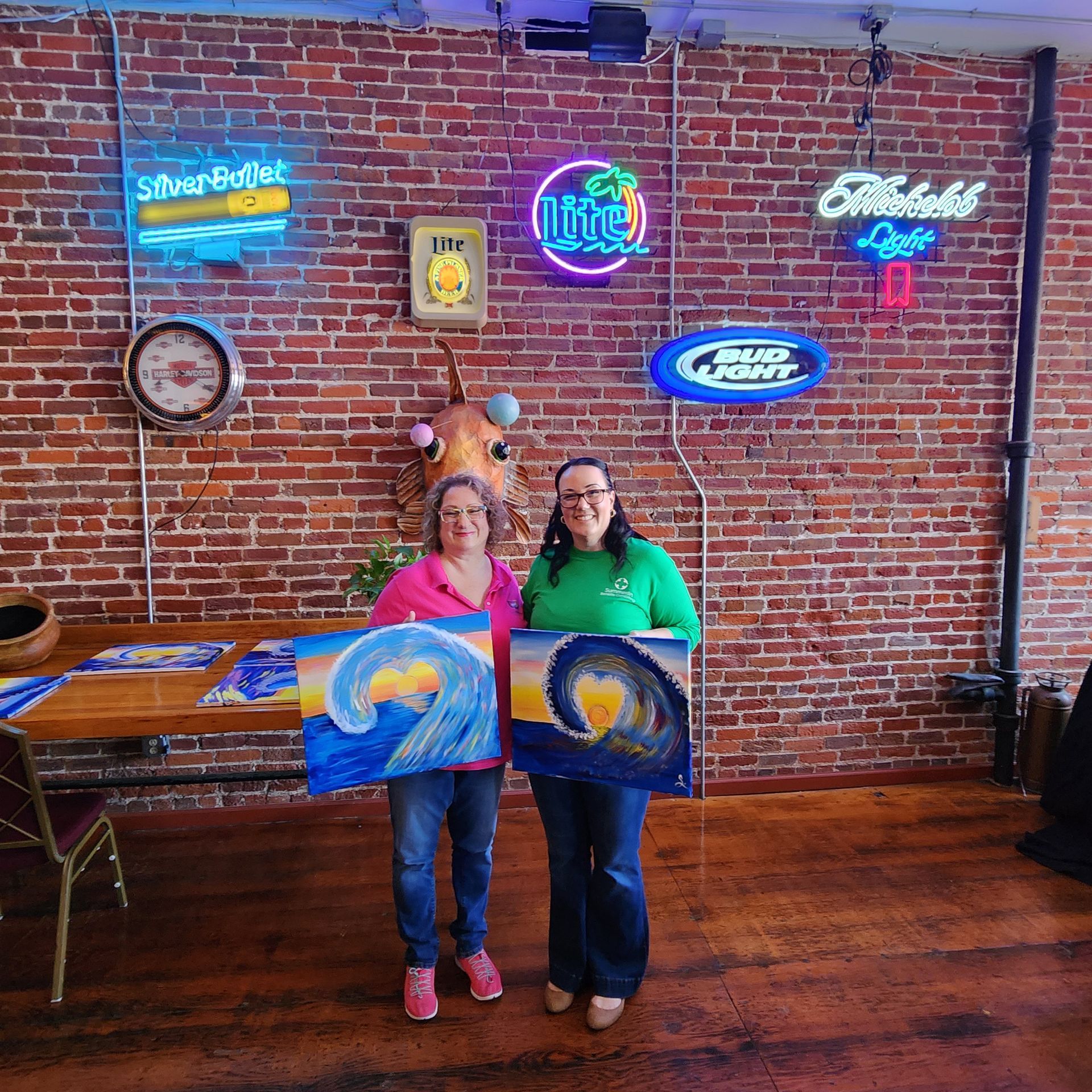 Two people holding paintings in a brick-walled venue with neon signs and a wooden floor.