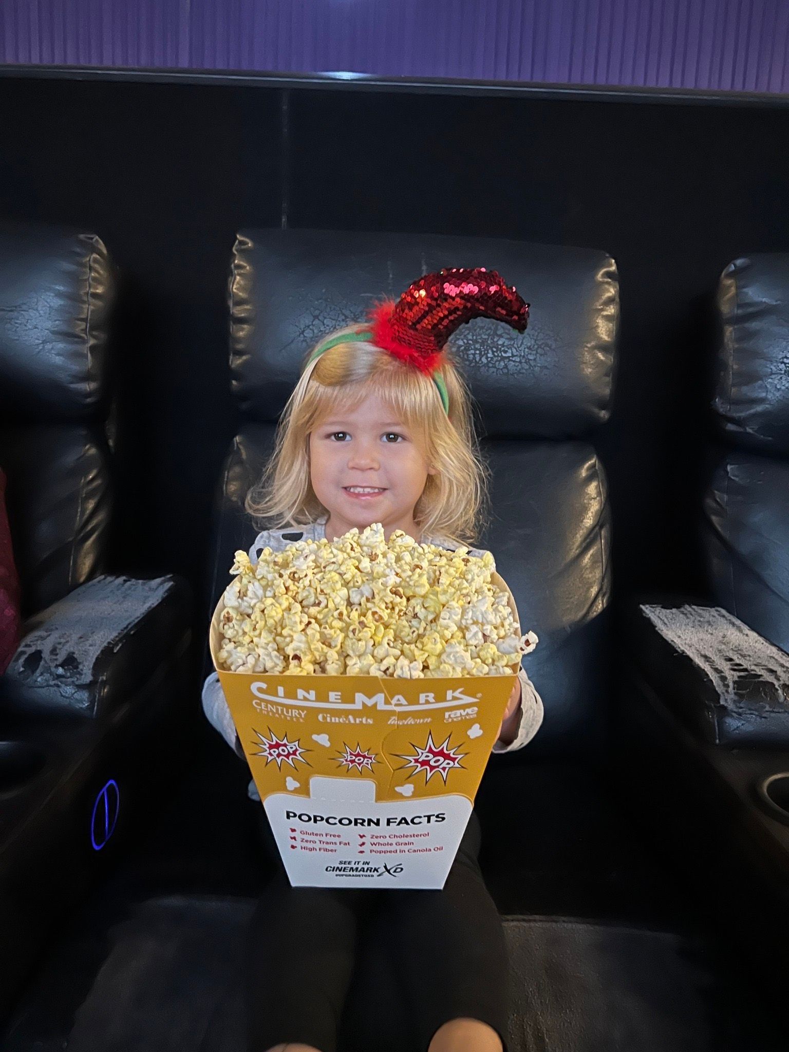 A little girl is sitting in a chair holding a box of popcorn.