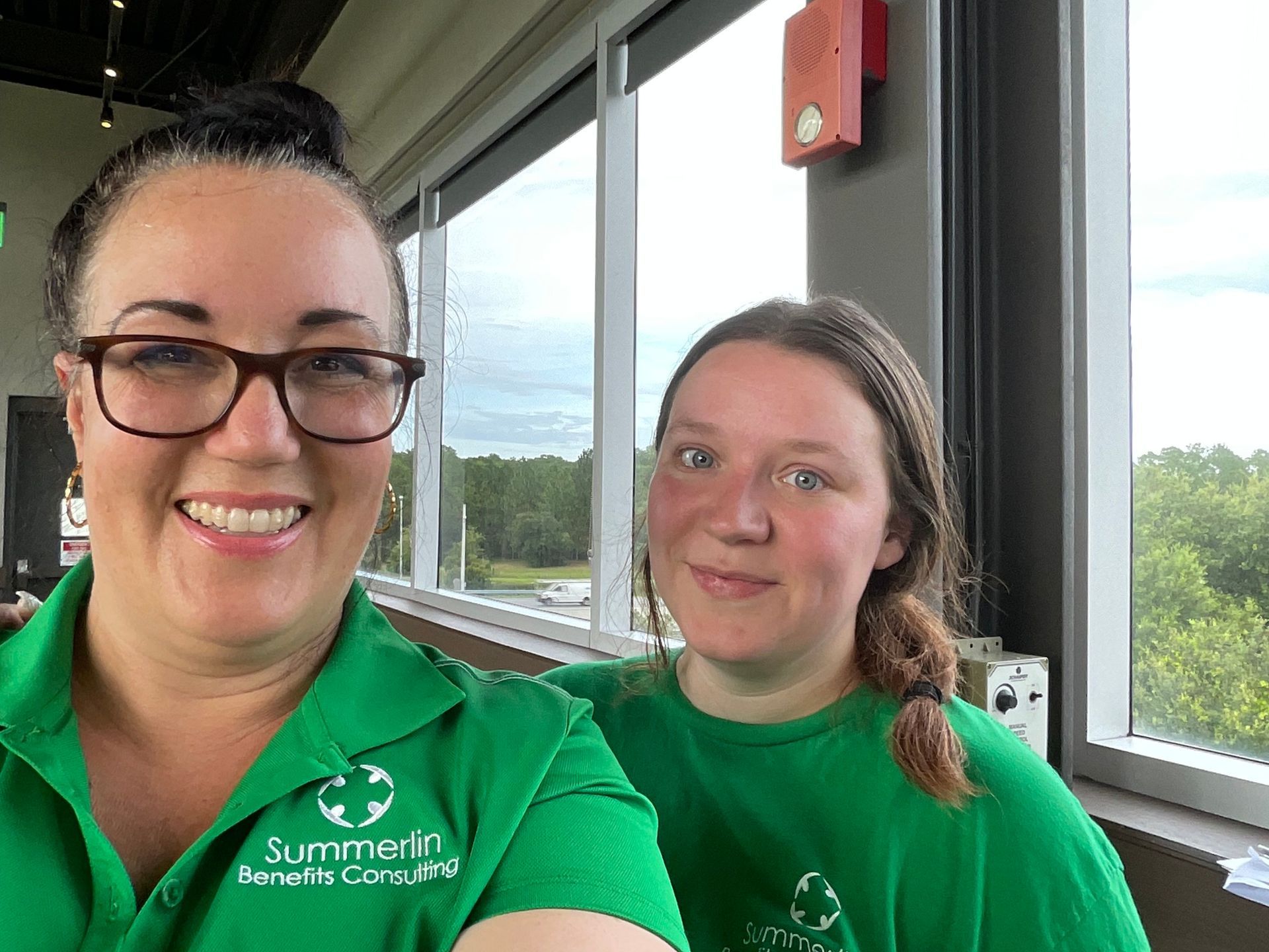 Two women in green shirts are taking a selfie in front of a window.