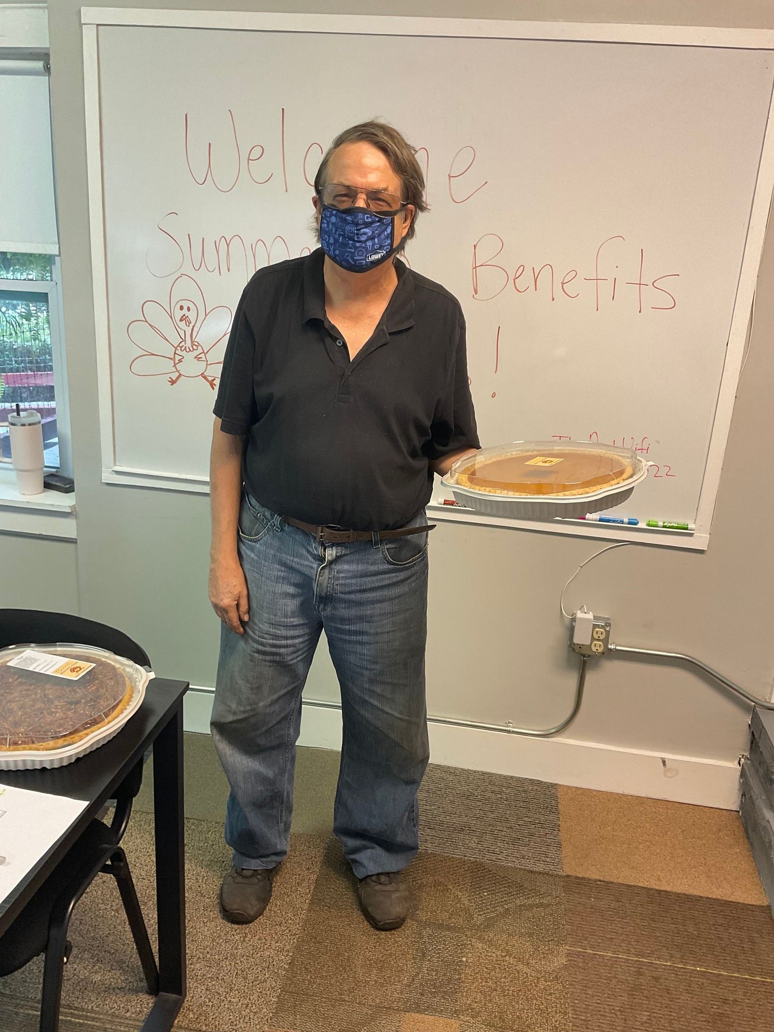 A man wearing a mask is holding a pie in front of a whiteboard.