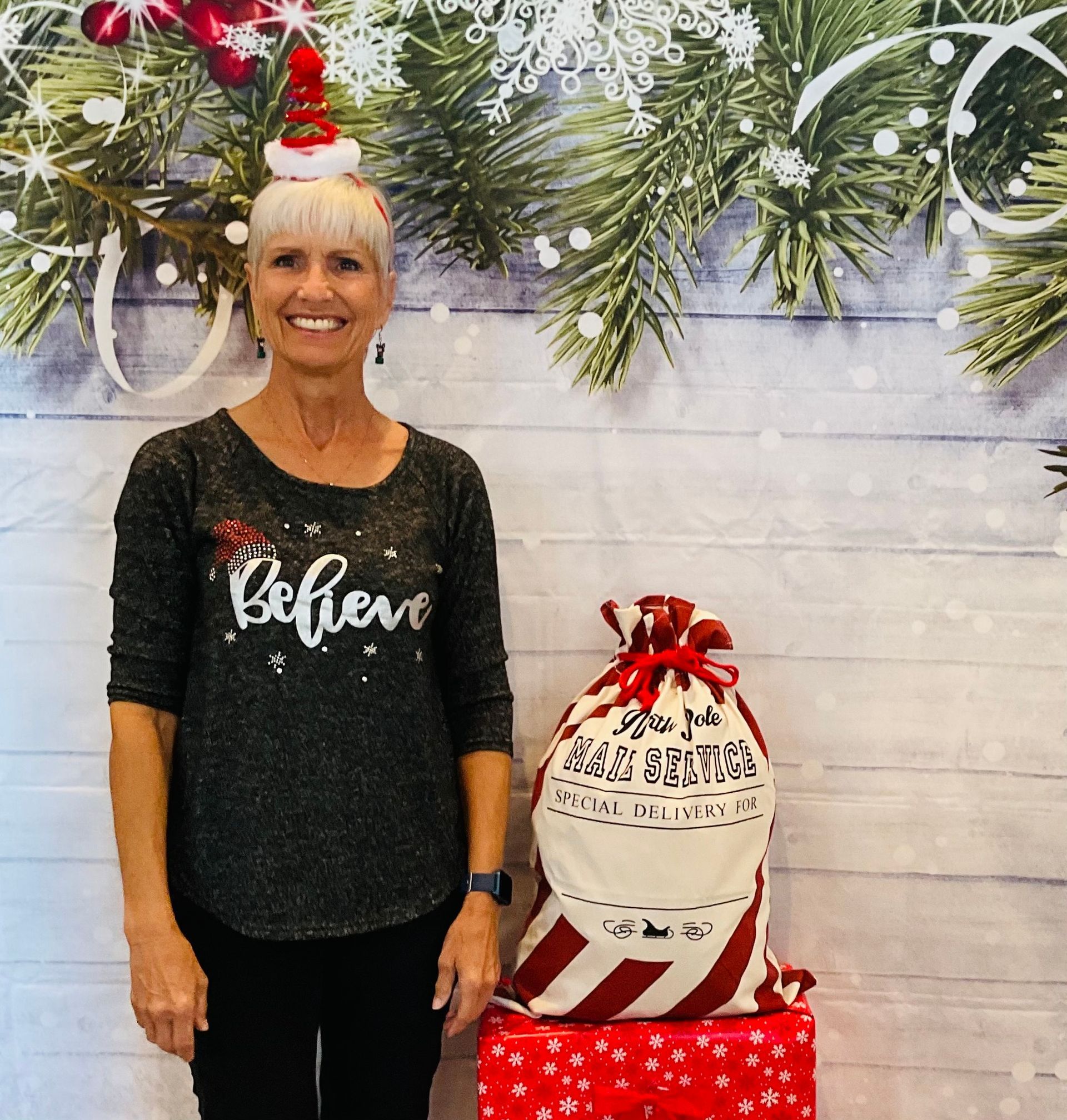 A woman wearing a sweater that says believe is standing in front of a christmas backdrop.