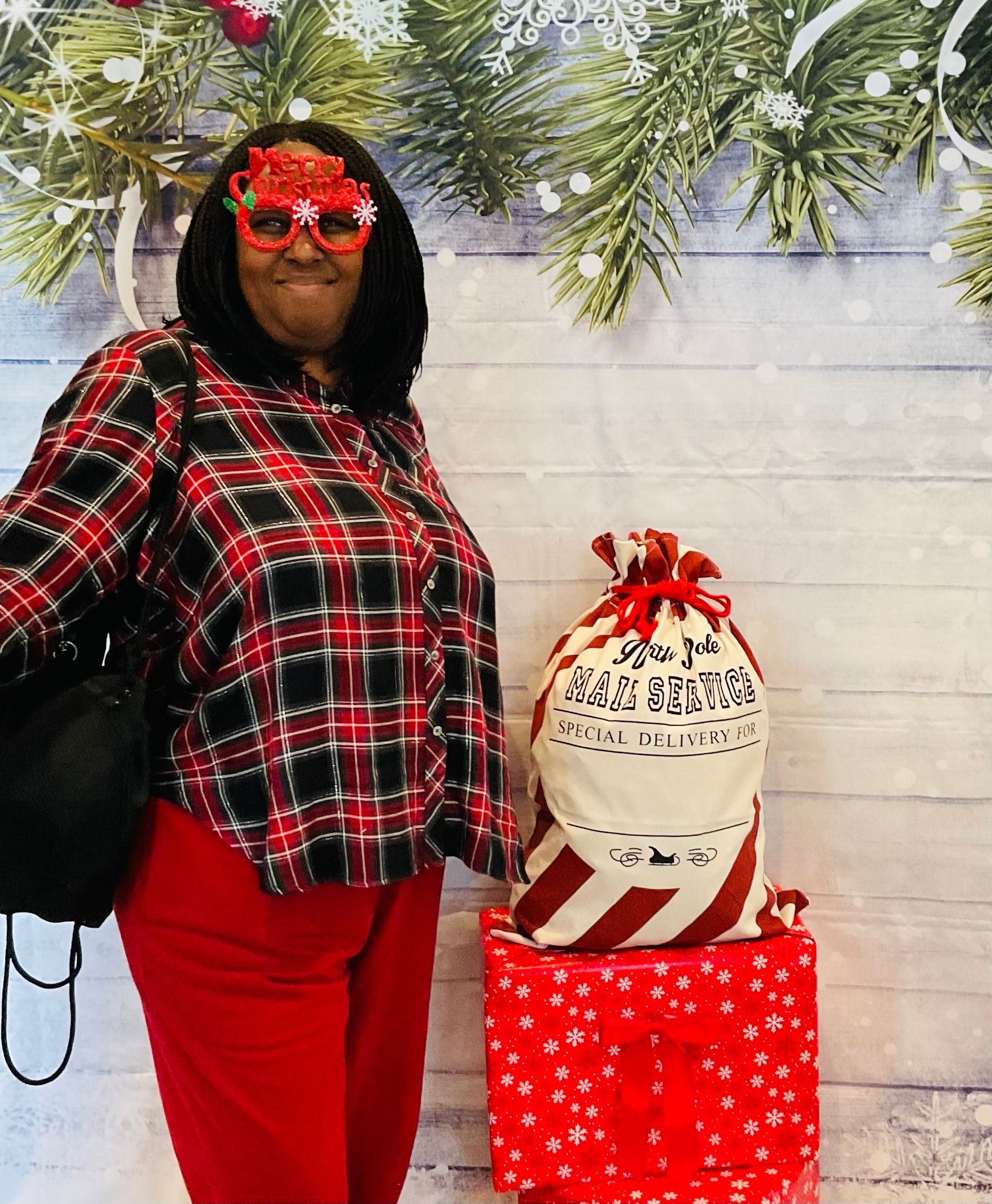 A woman wearing a plaid shirt and red pants is standing in front of a christmas background.