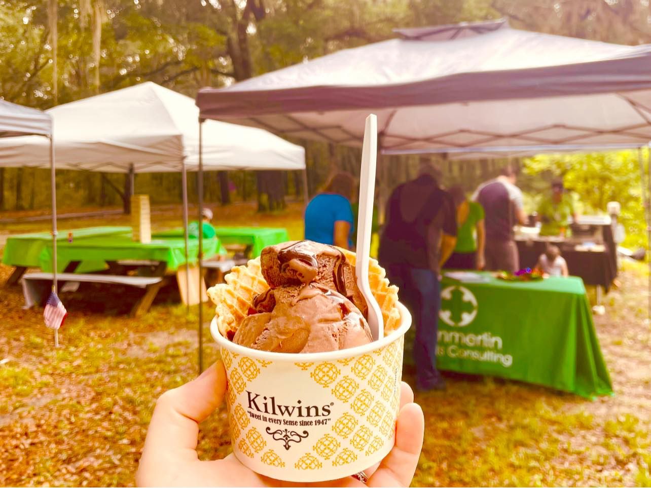 A person is holding a cup of chocolate ice cream in front of a tent.