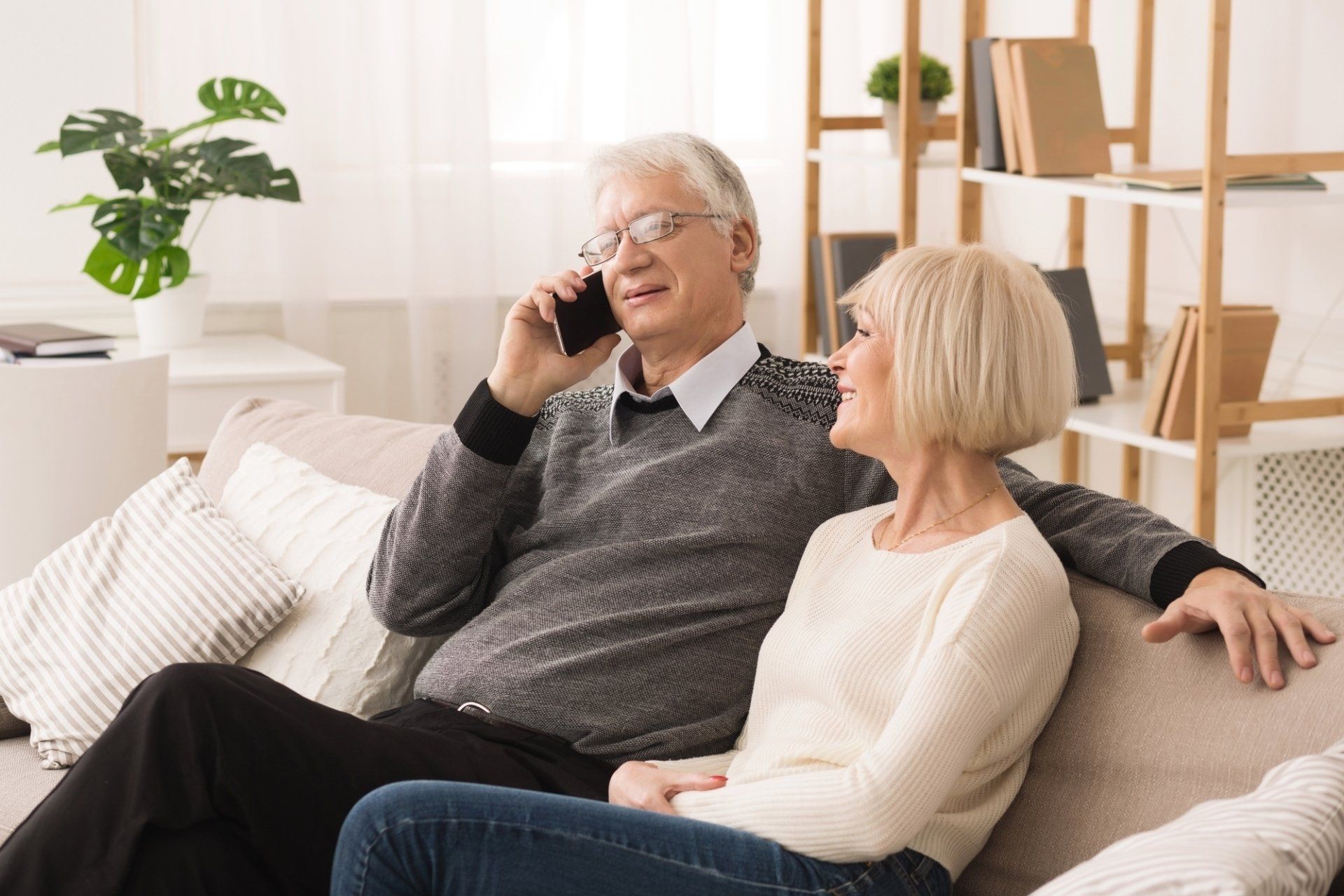 An elderly couple is sitting on a couch talking on a cell phone.