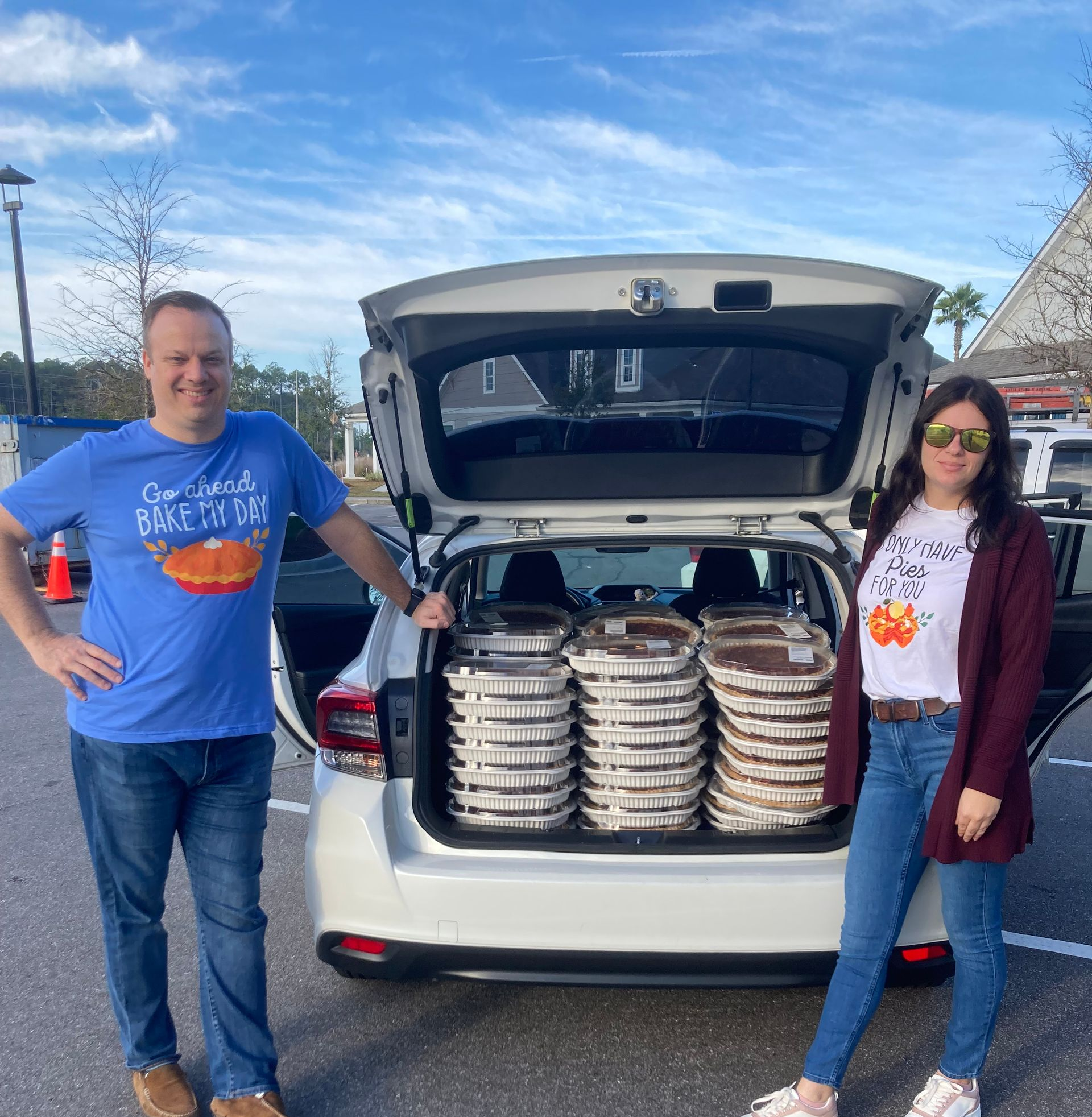 Two people stand beside an open car trunk filled with stacked food trays in a parking lot.