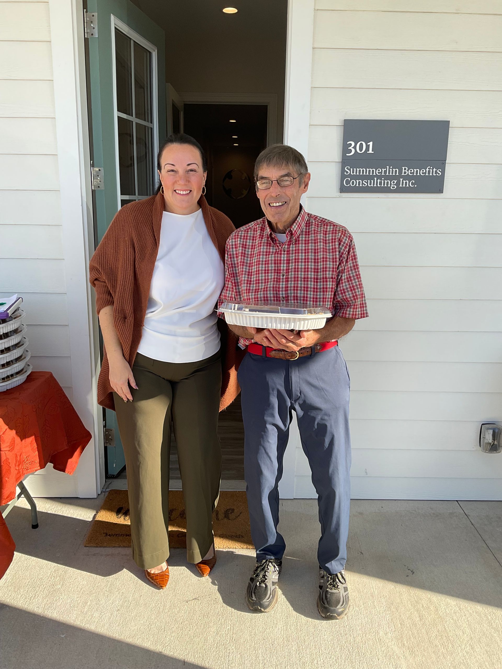 A man and a woman are standing in front of a building holding a tray of food.
