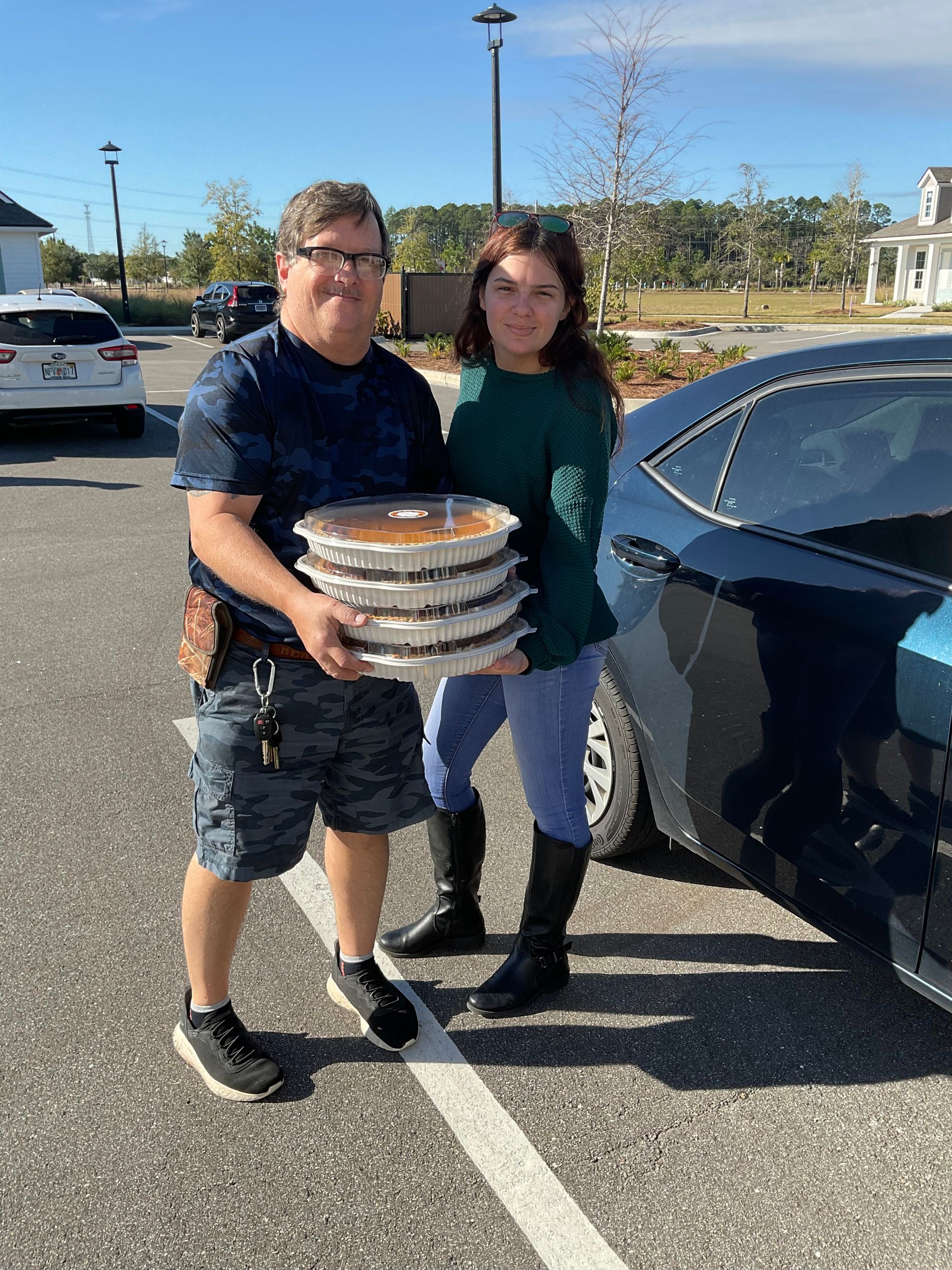 A man and a woman are holding a stack of pies in a parking lot.