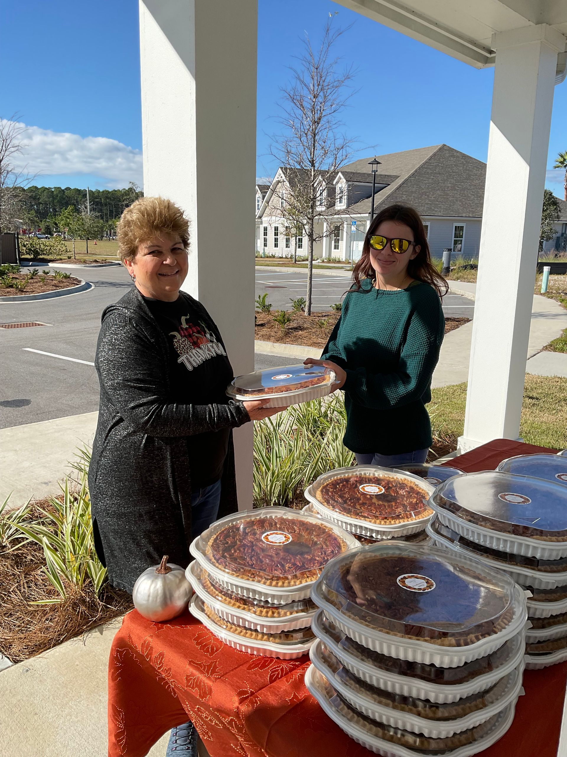 Two women are standing next to a table full of pies.