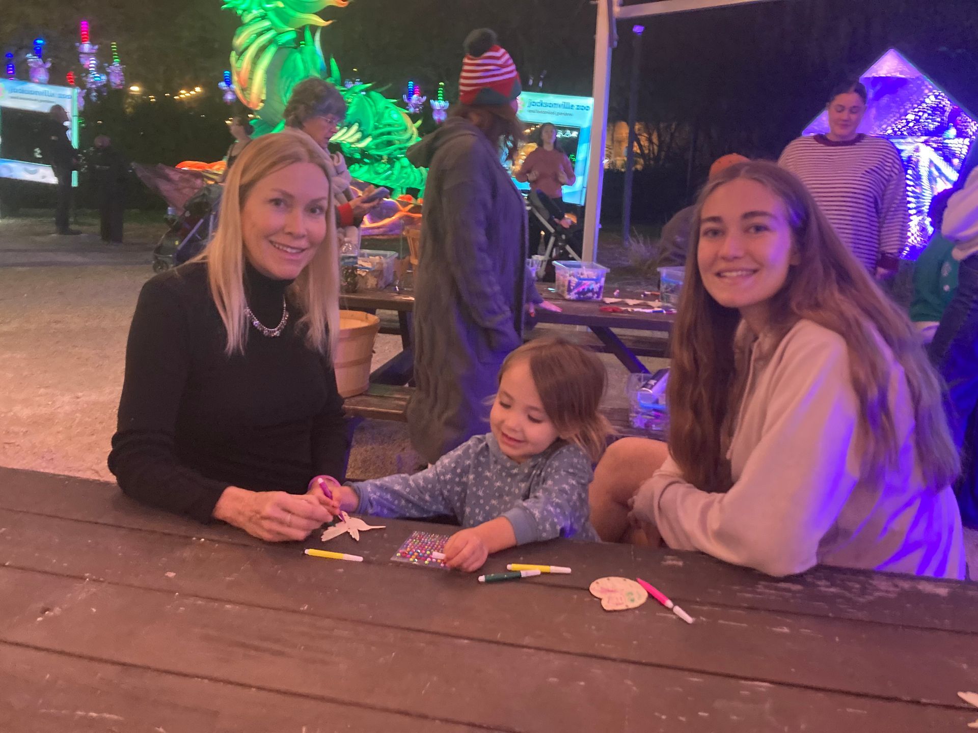 Three people smiling at a picnic table in a festive outdoor light display at night