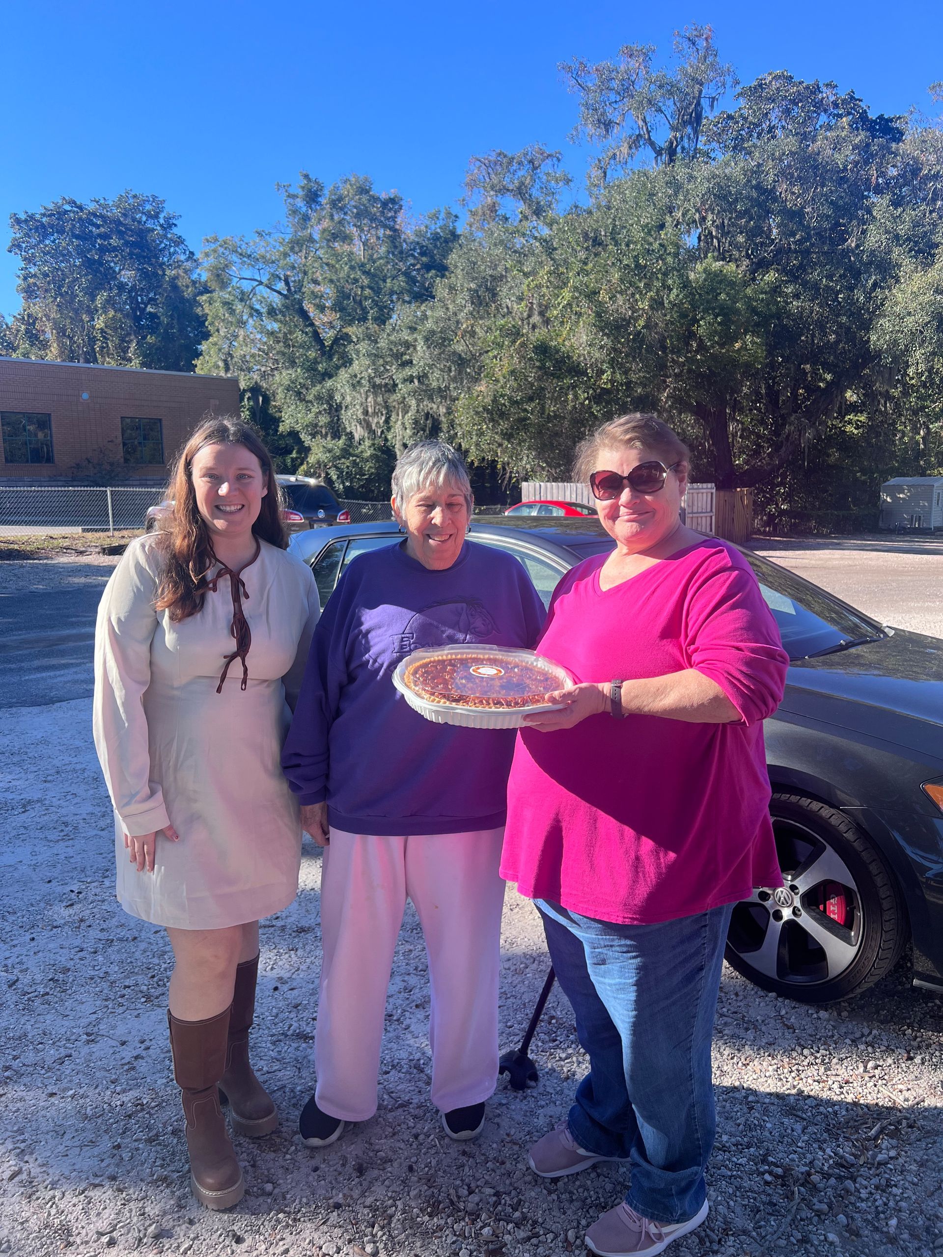Three women are standing next to each other in a parking lot holding a pie.