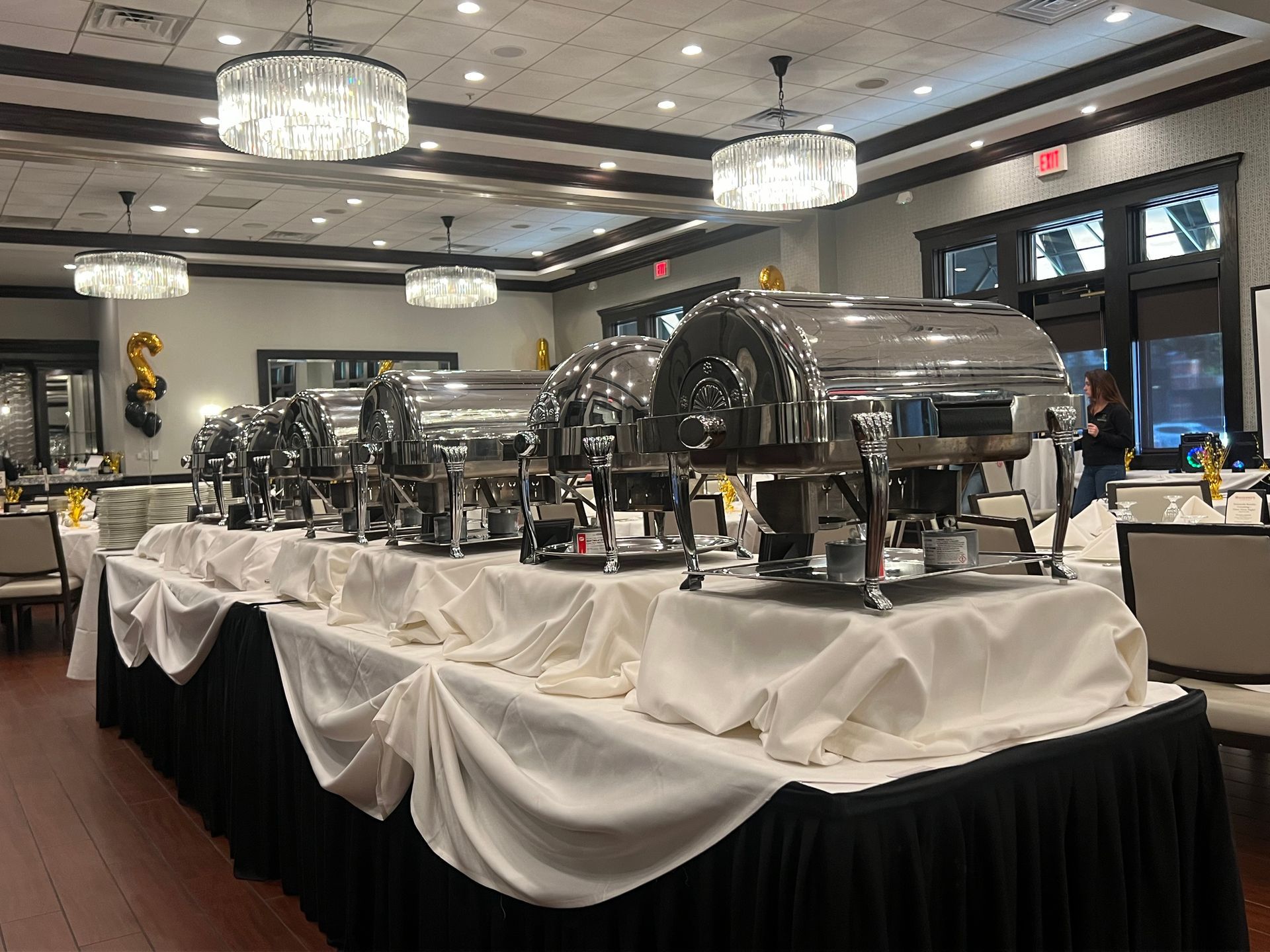 Buffet table set for an event with silver chafing dishes and white and black linens in a banquet hall.
