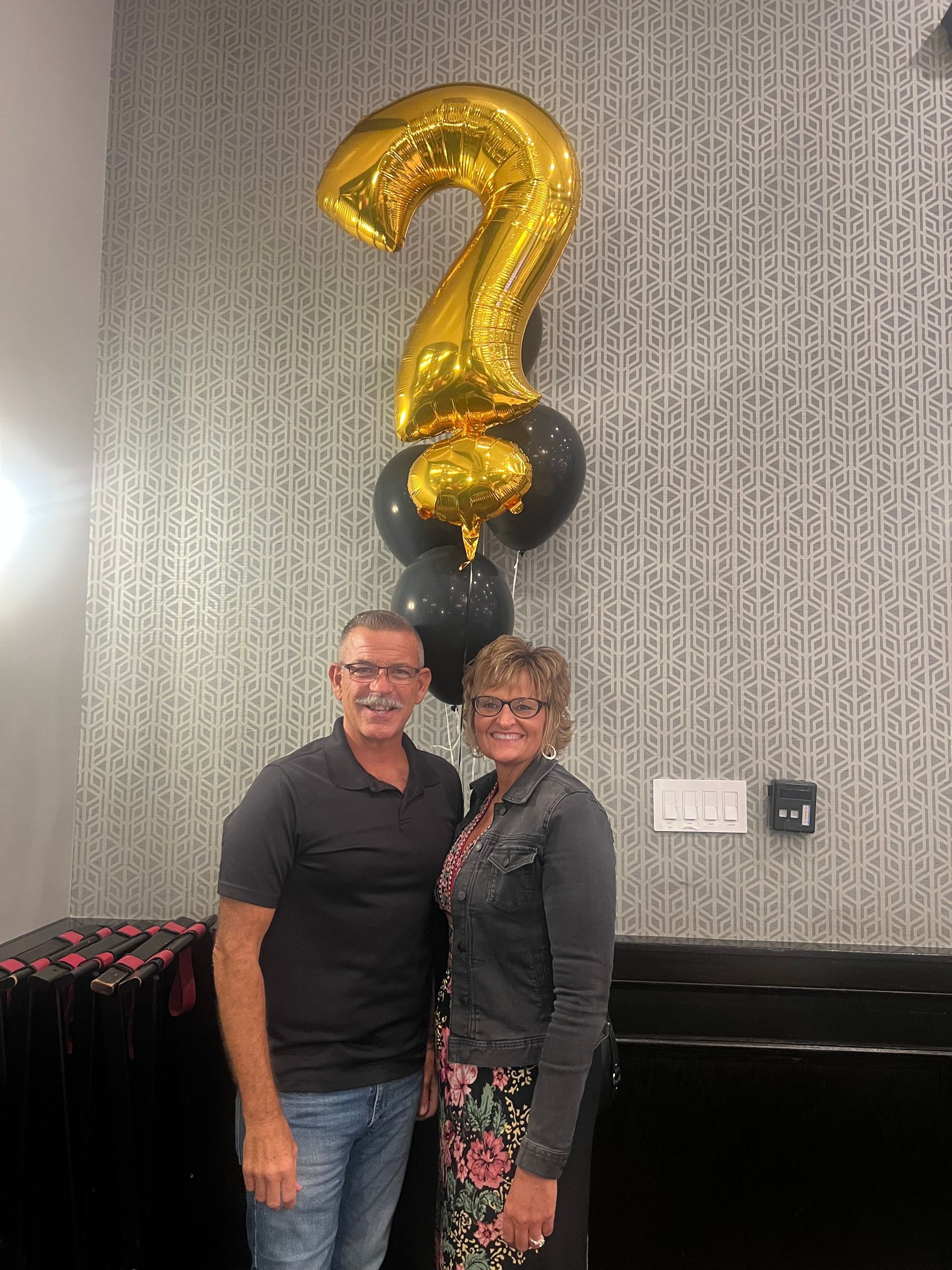 Couple standing, smiling, with a gold question mark balloon and black balloons in front of a patterned wall.