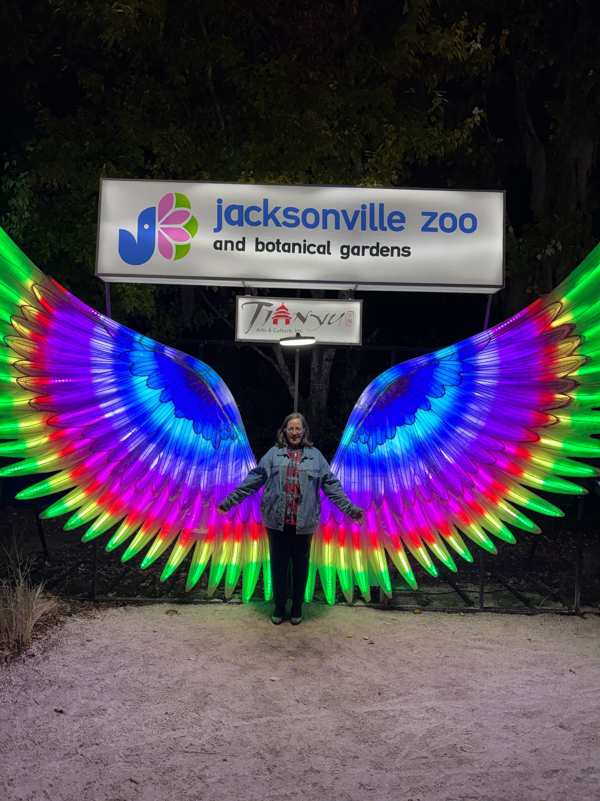 Person posing under Jacksonville Zoo sign with large rainbow-lit wings at night