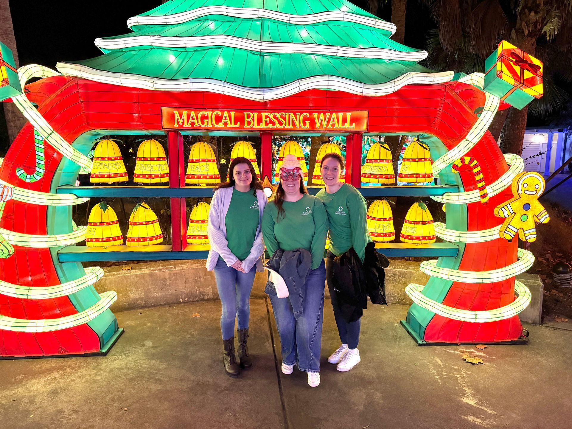 Three people posing in front of a colorful illuminated festive display with a red sign and lanterns