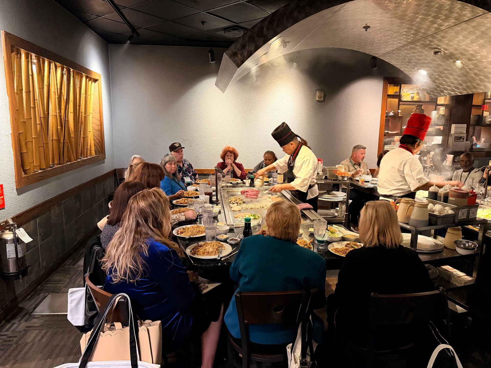 People dining at a long table in a restaurant with chefs serving food in the background