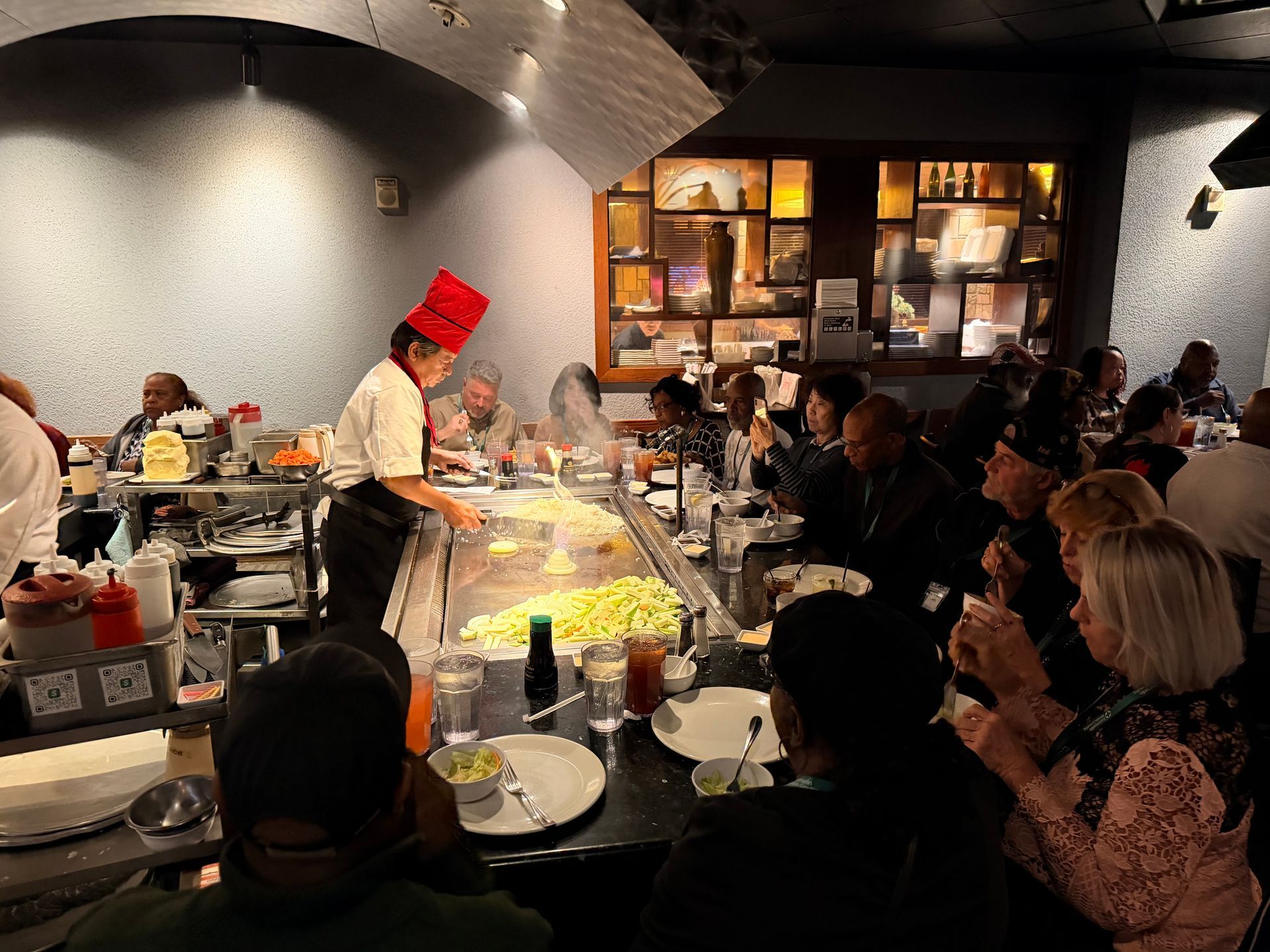 Chef in red hat cooks at a teppanyaki grill while diners watch in a busy restaurant