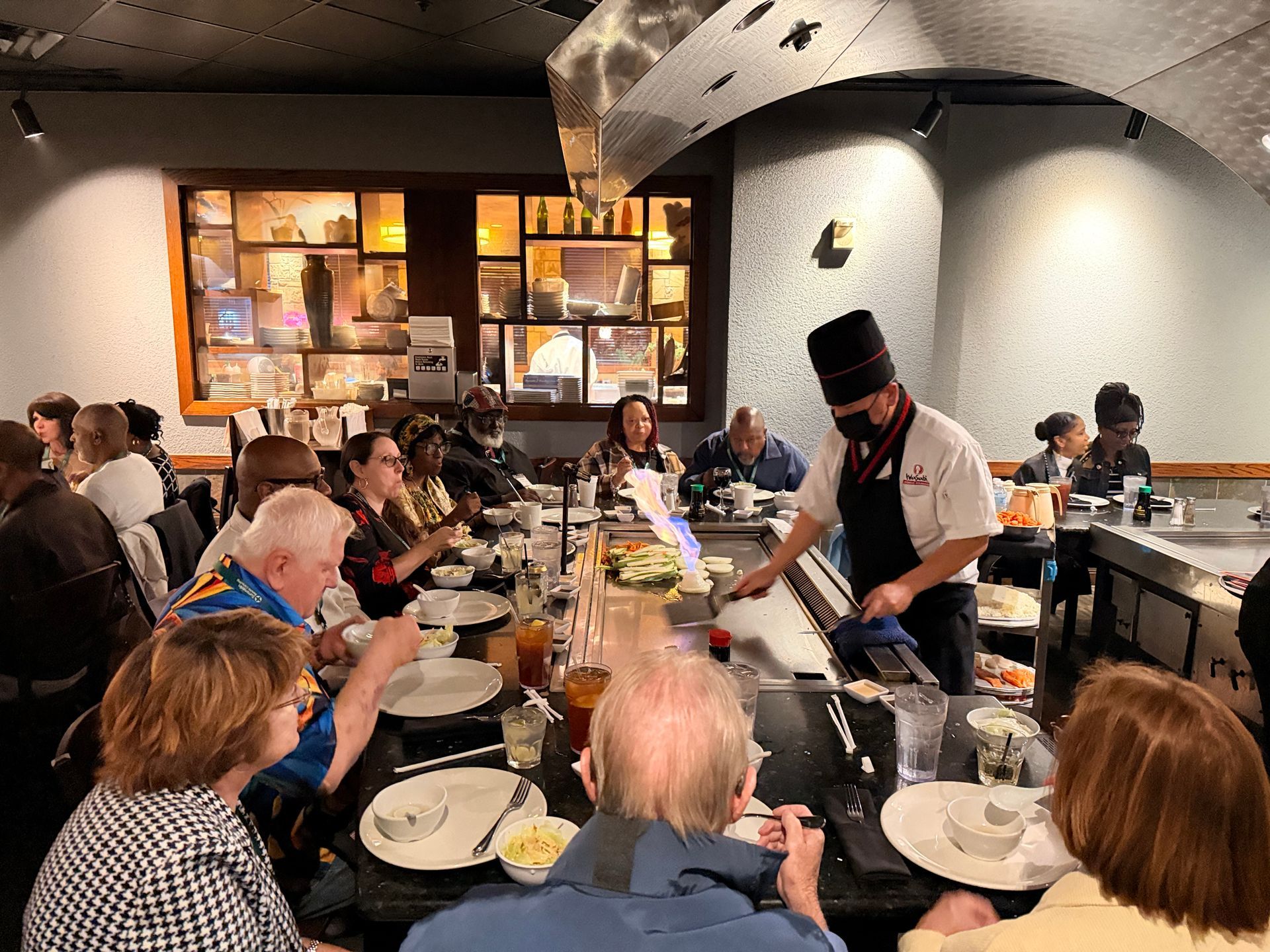 People seated at a counter watching a chef cook in a busy restaurant kitchen