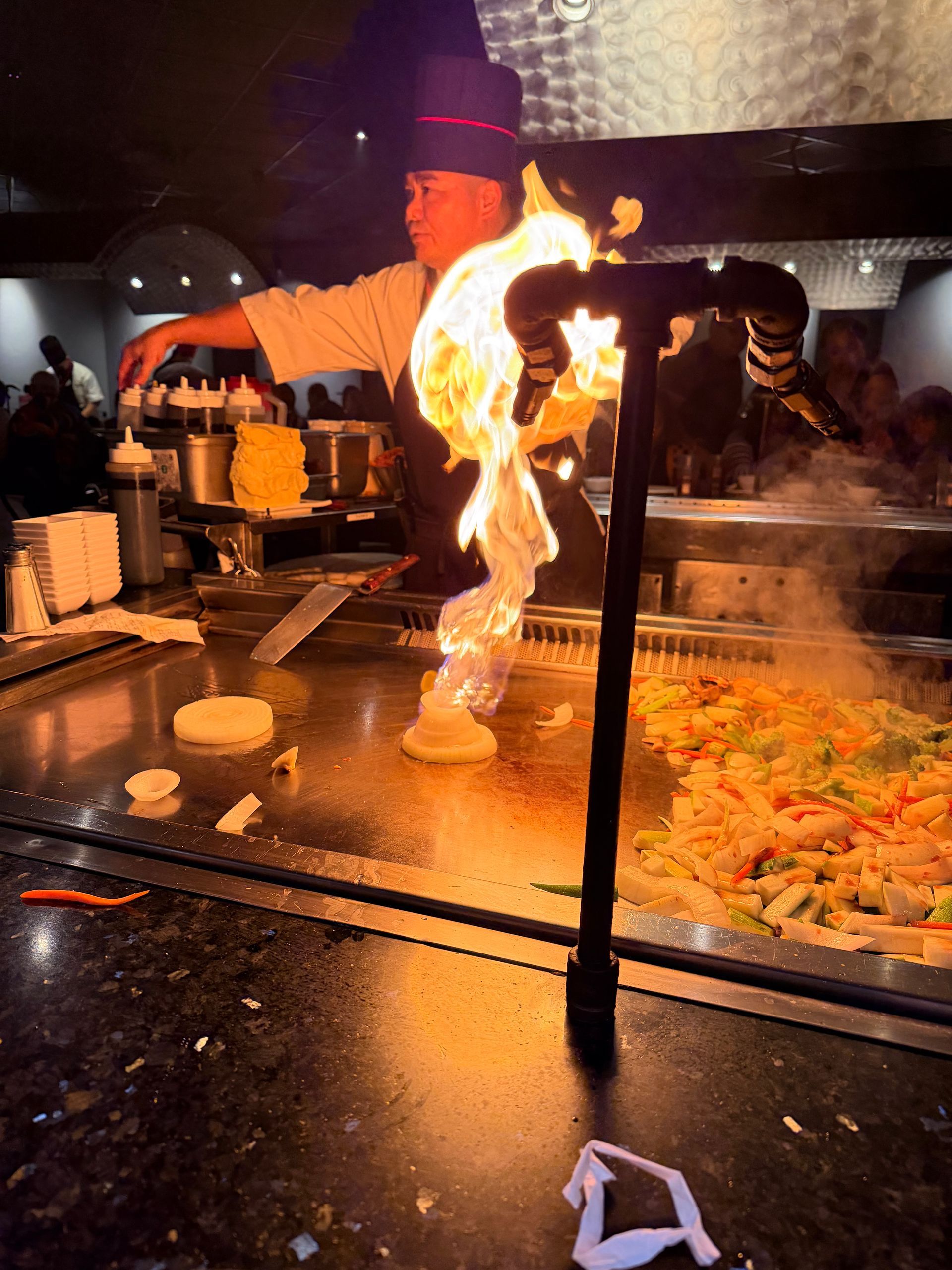 Chef cooking at a hibachi grill with a large flame, diners watching nearby