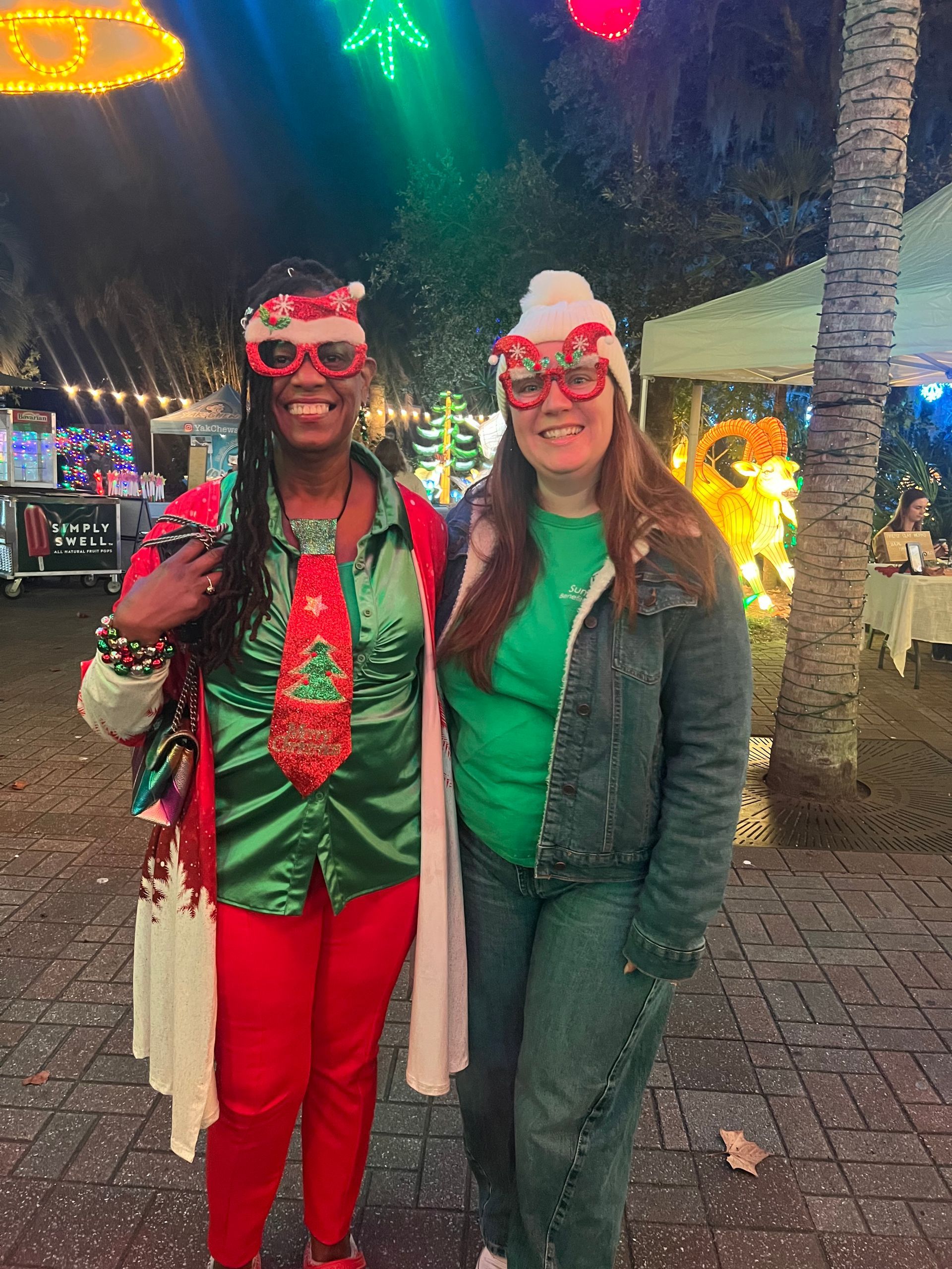 Two smiling people in festive red-and-green outfits and reindeer glasses standing outdoors under colorful lights.