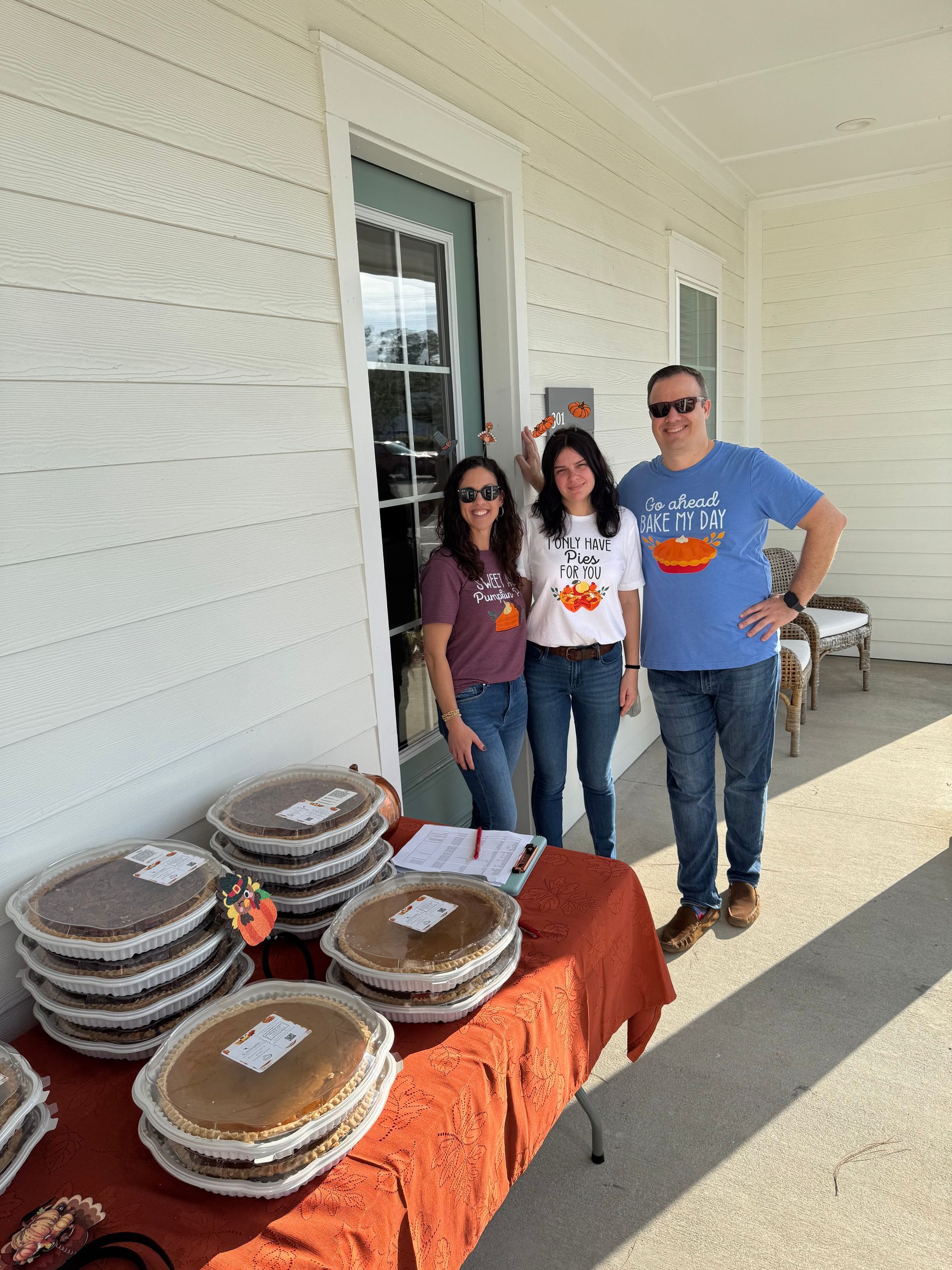 Three people standing beside a table of baked goods outside a white building.