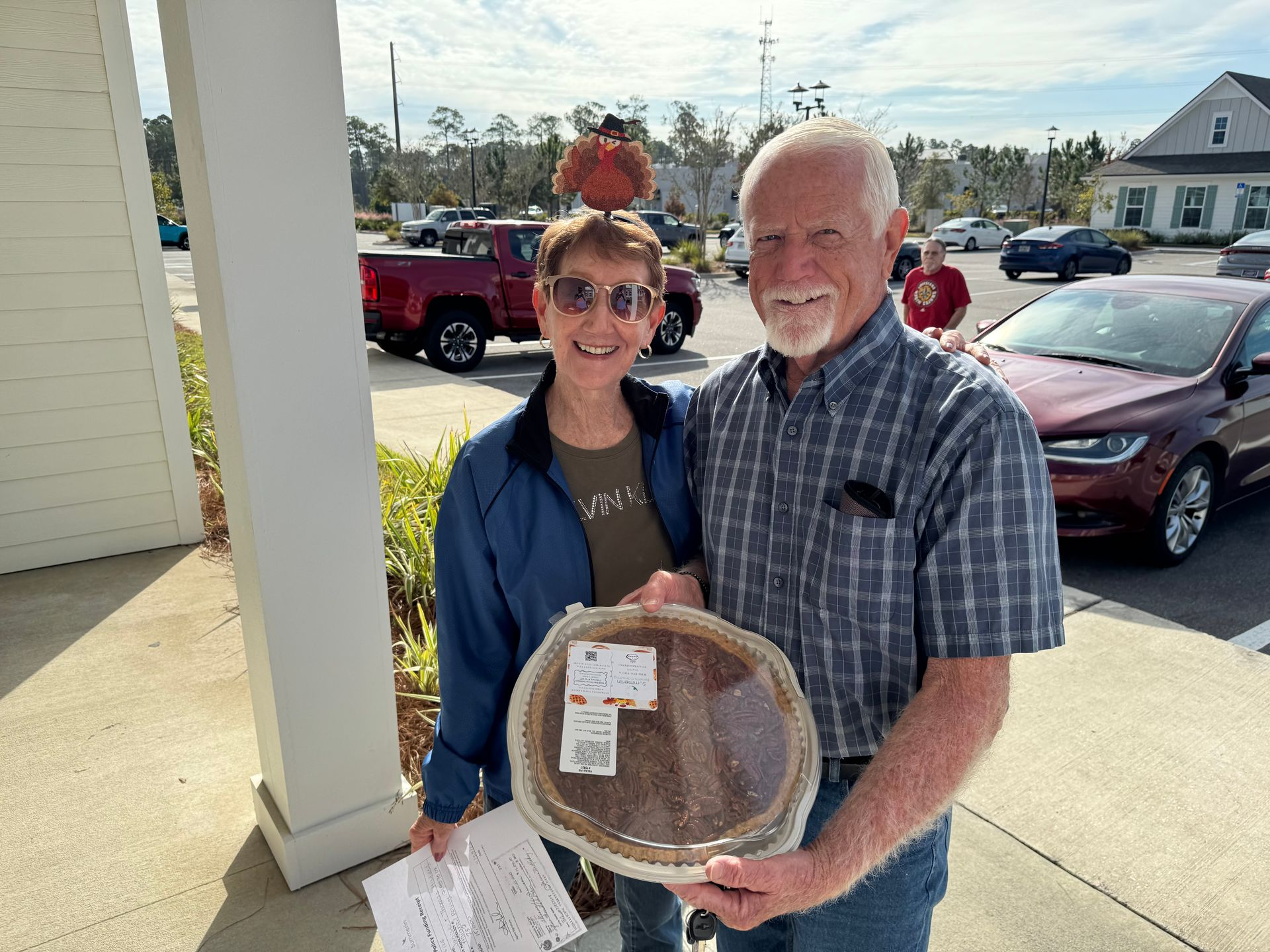 Two smiling people outdoors holding a packaged pie in a parking lot