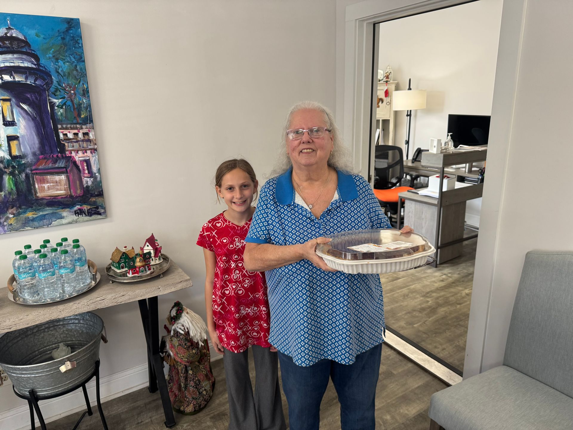 Woman and girl holding a cake in a living room beside a table with desserts
