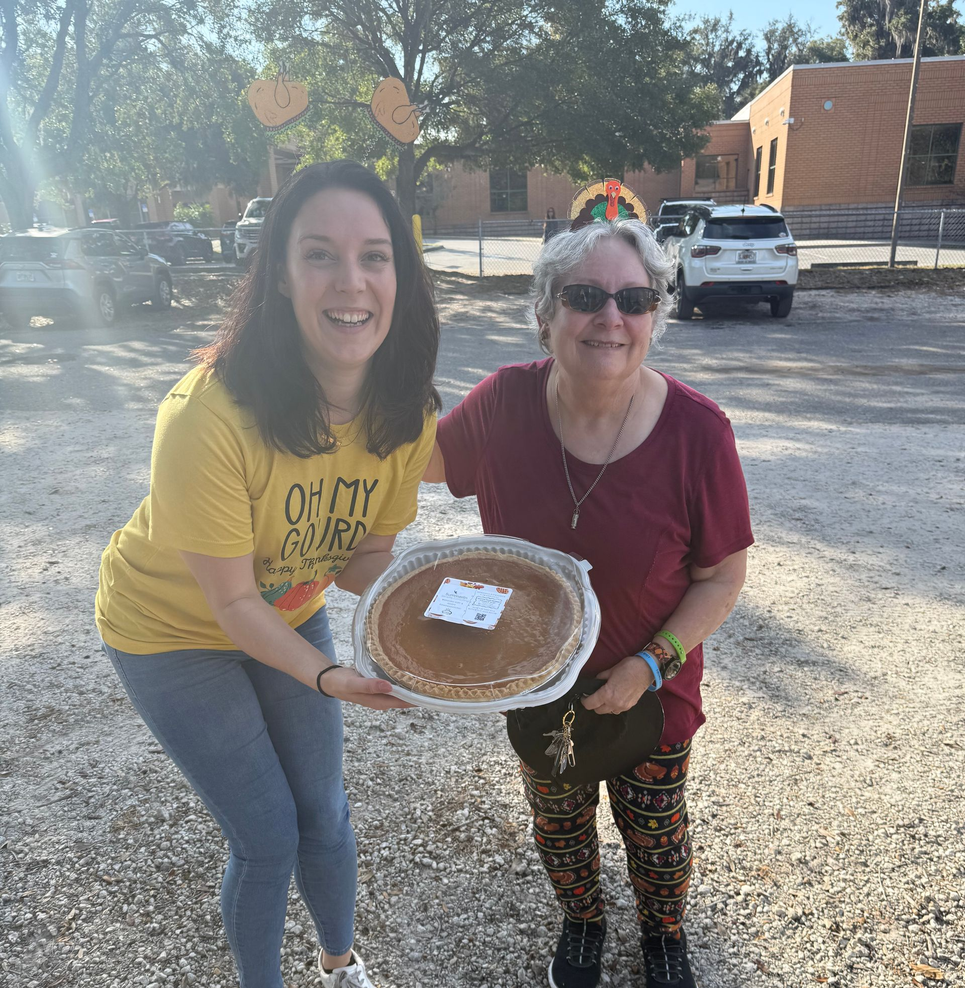 Two smiling people holding a pie outdoors in a sunny parking lot